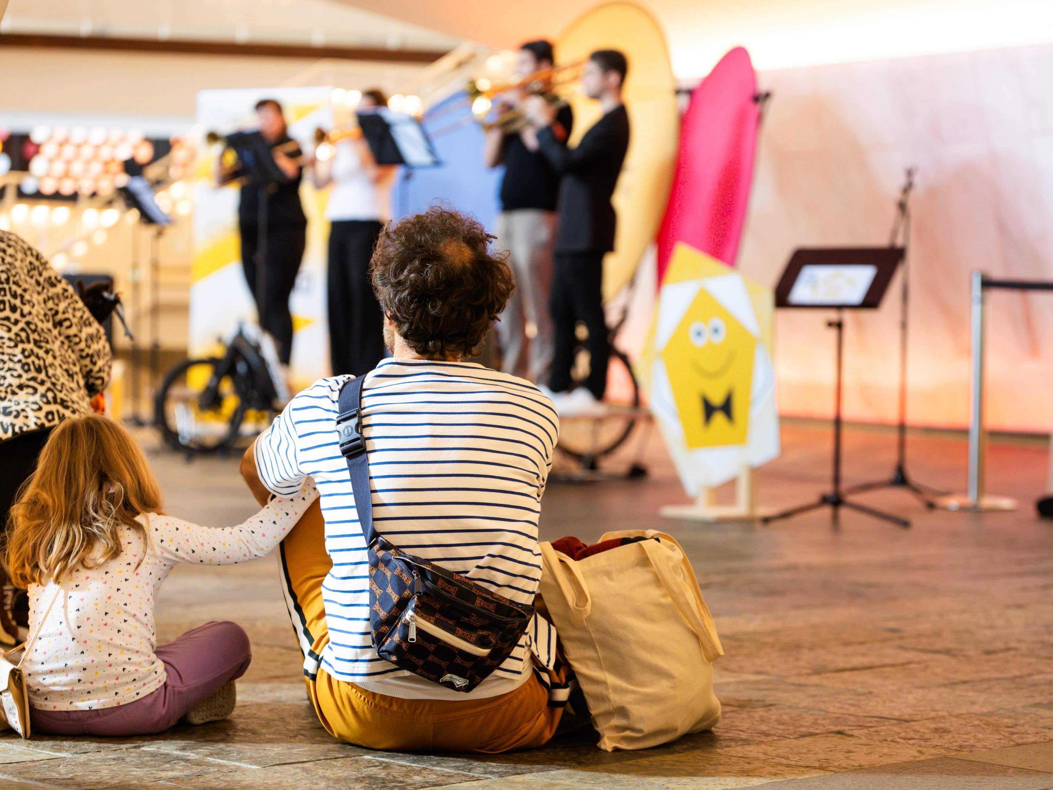Ein Mann und ein Kind sitzen auf dem Boden und sehen zu, wie ein Blechbläserensemble auf der VeloStage spielt mit Notenständern im Hintergrund.