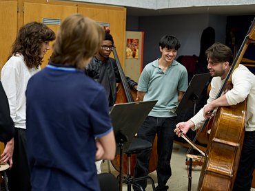 Eine Gruppe von Schülern steht im Kreis, einige halten Kontrabässe in den Händen, während ein Lehrer an einem Kontrabass eine Technik vorführt.