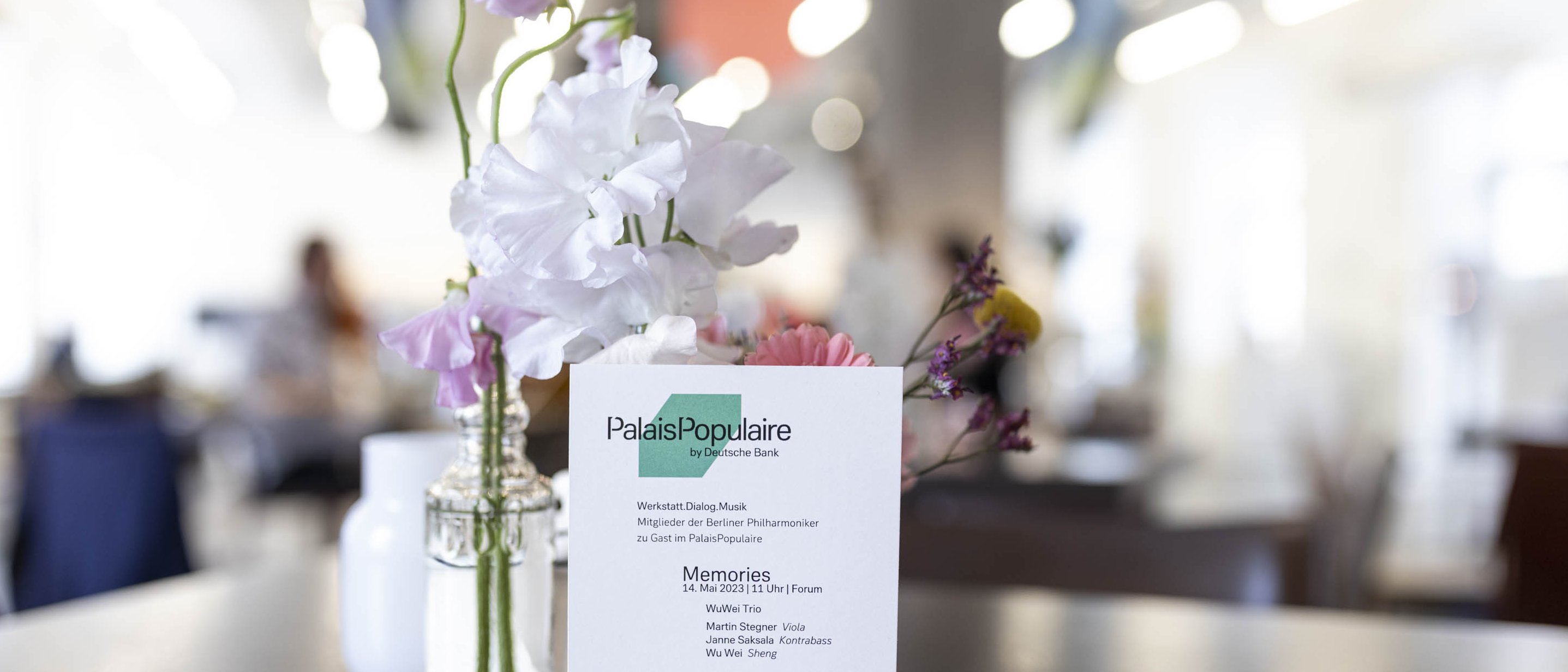 Auf einem Tisch in einem Café steht eine kleine Vase mit rosa und weißen Blumen neben einer Broschüre oder einem Menü für PalaisPopulaire, auf dem eine Veranstaltung der Berliner Philharmoniker vorgestellt wird.