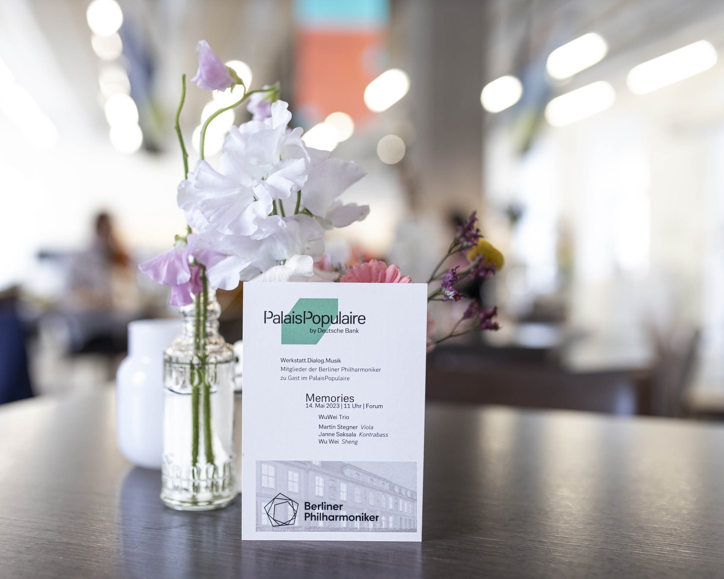 Auf einem Tisch in einem Café steht eine kleine Vase mit rosa und weißen Blumen neben einer Broschüre oder einem Menü für PalaisPopulaire, auf dem eine Veranstaltung der Berliner Philharmoniker vorgestellt wird.