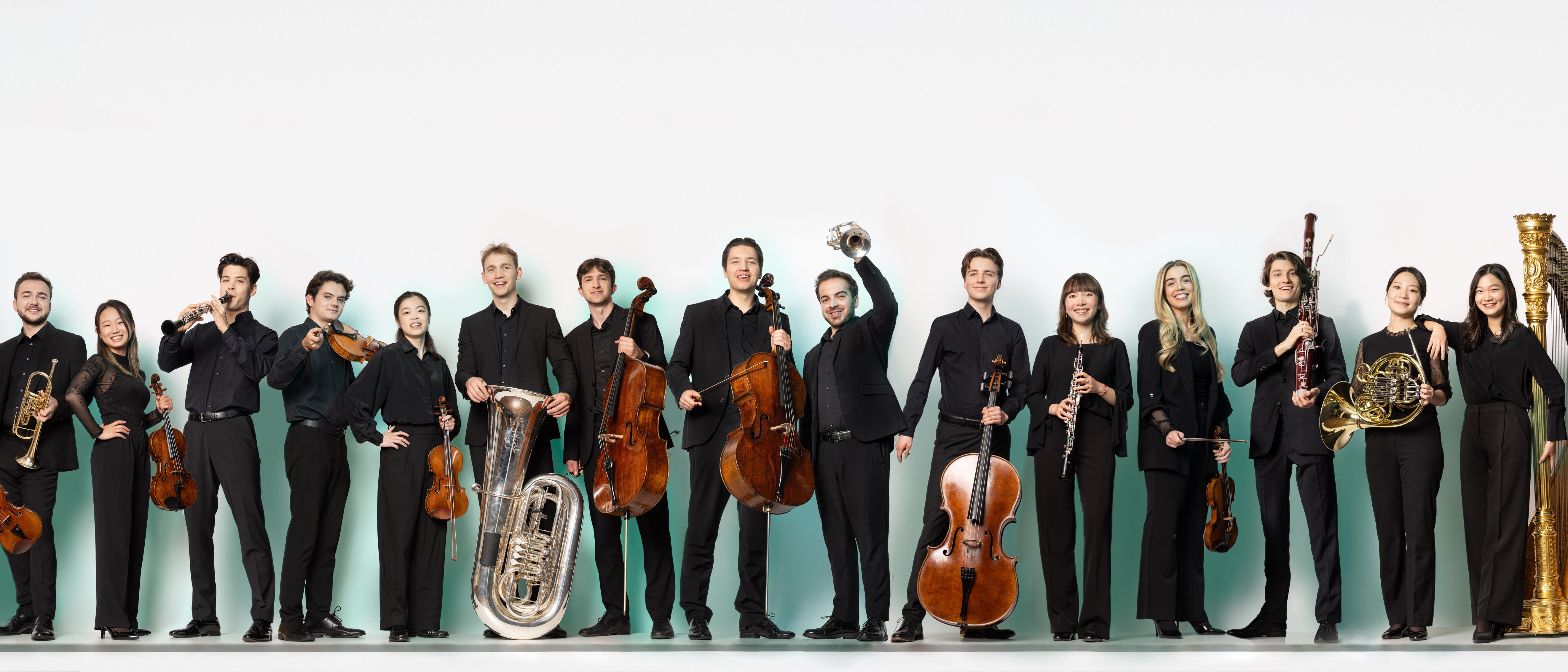 Mitglieder der Karajan-Akademie in formeller schwarzer Kleidung steht in einer Reihe, jeder hält sein Musikinstrument, darunter Streicher, Blechbläser, Holzbläser und Schlagzeug, vor einem schlichten weißen Hintergrund.