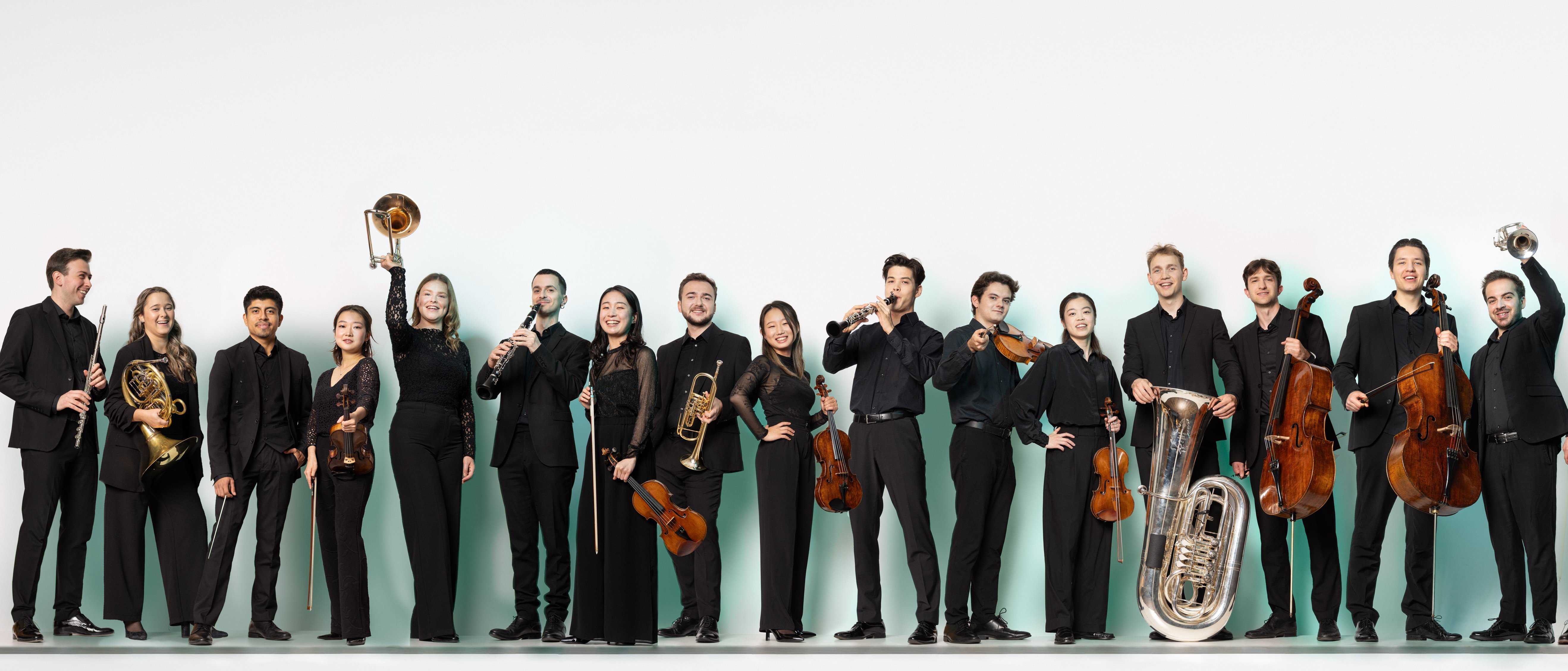 Mitglieder der Karajan-Akademie in formeller schwarzer Kleidung steht in einer Reihe, jeder hält sein Musikinstrument, darunter Streicher, Blechbläser, Holzbläser und Schlagzeug, vor einem schlichten weißen Hintergrund.