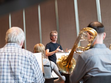 Raphael Haeger in einem schwarzen Hemd leitet eine Orchesterprobe. Die Musiker, einige mit Blechblasinstrumenten, sitzen in einem modernen, gut beleuchteten Raum mit hohen Fenstern und sind dem Dirigenten zugewandt.