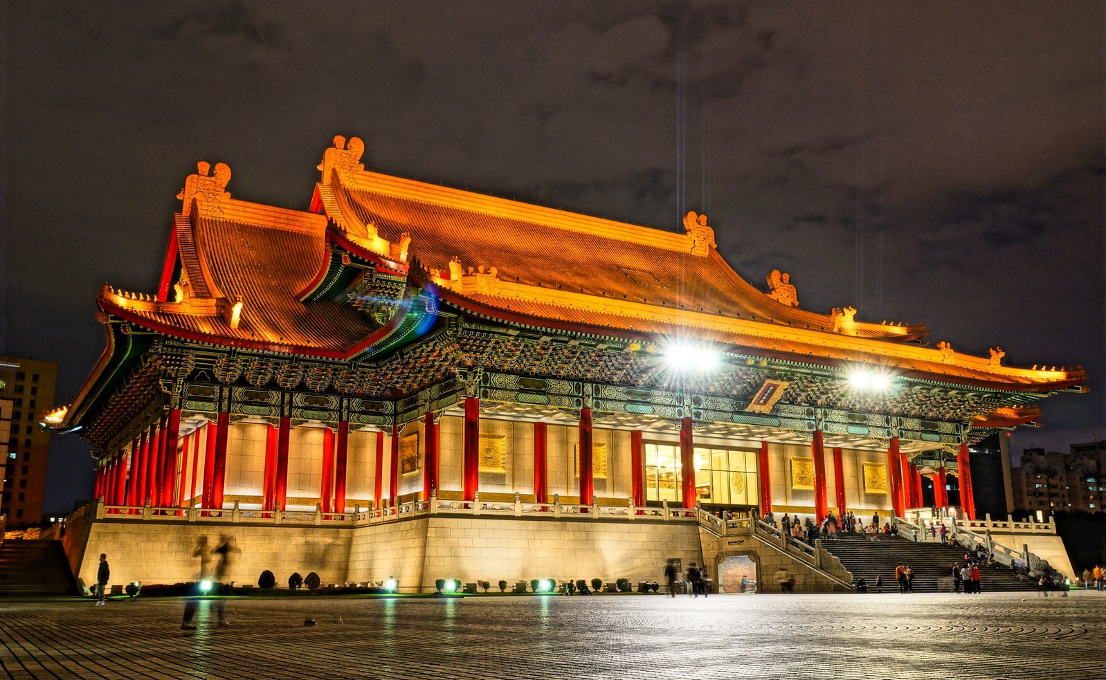 Ein hell erleuchtetes traditionelles chinesisches Gebäude mit verzierten roten Säulen und einem geschwungenen Dach steht vor einem nächtlichen Himmel, auf den Stufen und dem offenen Platz davor sind Menschen zu sehen.