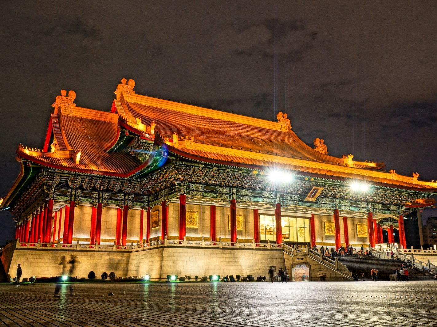 A large traditional Chinese building with ornate, illuminated roofs and red columns stands brightly lit at night, with a spacious stone plaza and people gathered near the entrance.