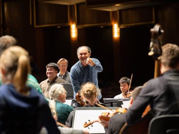 Ein Dirigent steht lächelnd und gestikulierend in Richtung eines Orchesters während einer Probe, die Musiker sitzen, halten ihre Instrumente und warmes Licht beleuchtet die Szene.