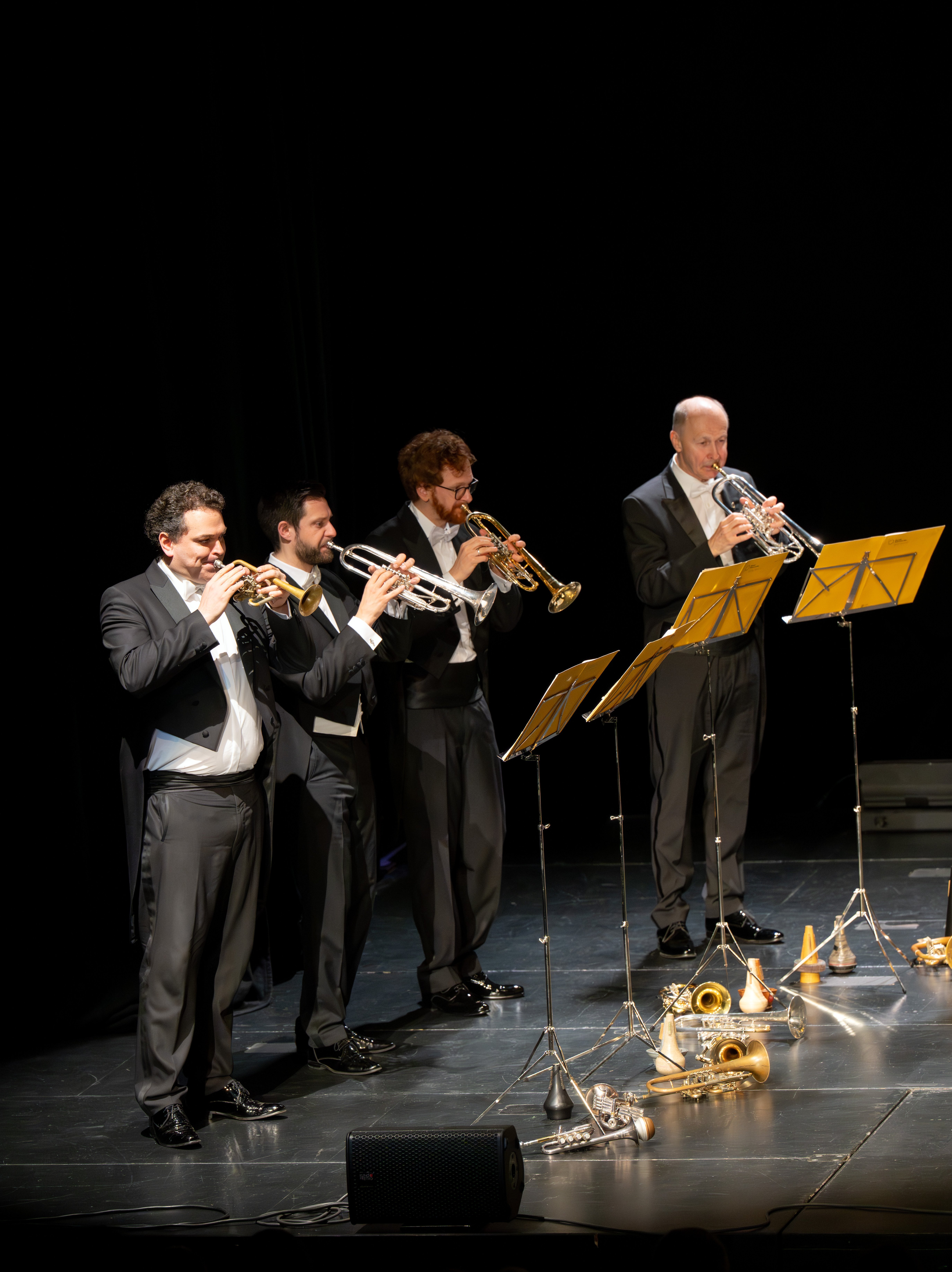 Vier Männer in formellen schwarzen Anzügen spielen Trompeten auf einer dunklen Bühne und stehen hinter gelben Notenständern. Vor ihnen auf dem Boden liegen mehrere Blechdämpfer und Instrumente.