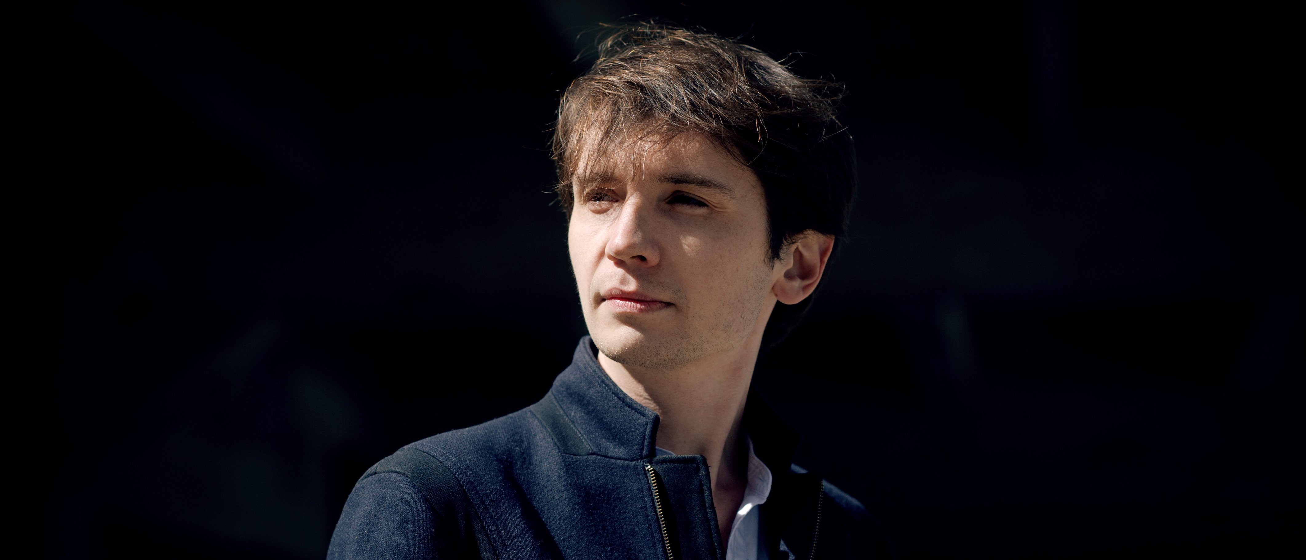 Karol Mossakowski mit braunem Haar, dunkler Jacke und weißeKarol Mossakowski, with brown hair, a dark jacket, and a white shirt, stands against a dark background, gazing thoughtfully to the side. The lighting highlights his face, creating a dramatic contrast.m Hemd steht vor einem dunklen Hintergrund und blickt nachdenklich zur Seite. Die Beleuchtung hebt sein Gesicht hervor und erzeugt einen dramatischen Kontrast.