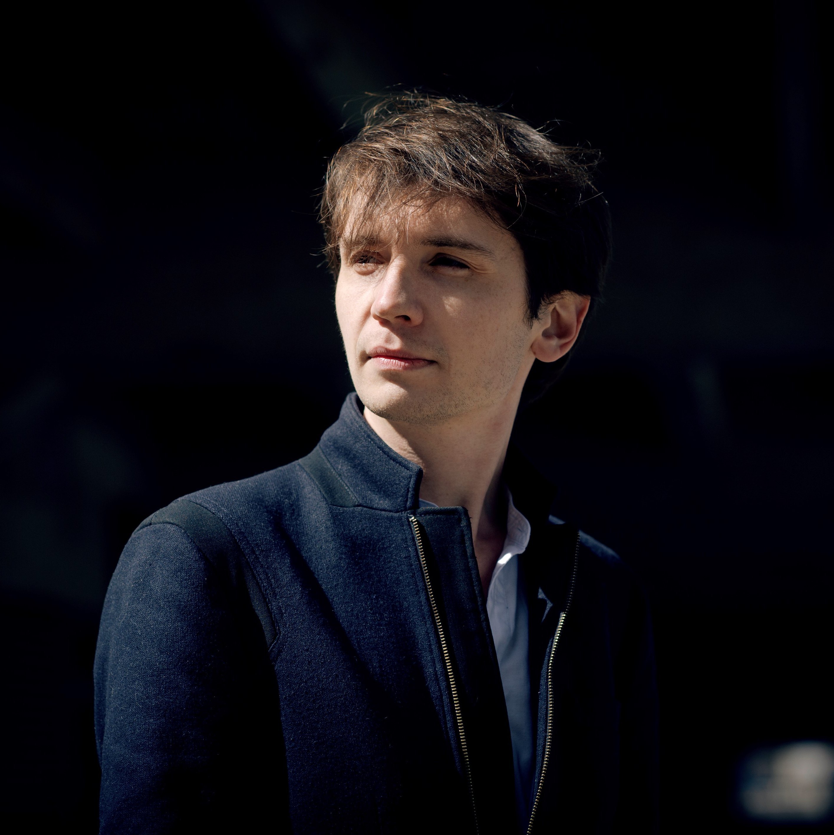 Karol Mossakowski mit braunem Haar, dunkler Jacke und weißeKarol Mossakowski, with brown hair, a dark jacket, and a white shirt, stands against a dark background, gazing thoughtfully to the side. The lighting highlights his face, creating a dramatic contrast.m Hemd steht vor einem dunklen Hintergrund und blickt nachdenklich zur Seite. Die Beleuchtung hebt sein Gesicht hervor und erzeugt einen dramatischen Kontrast.