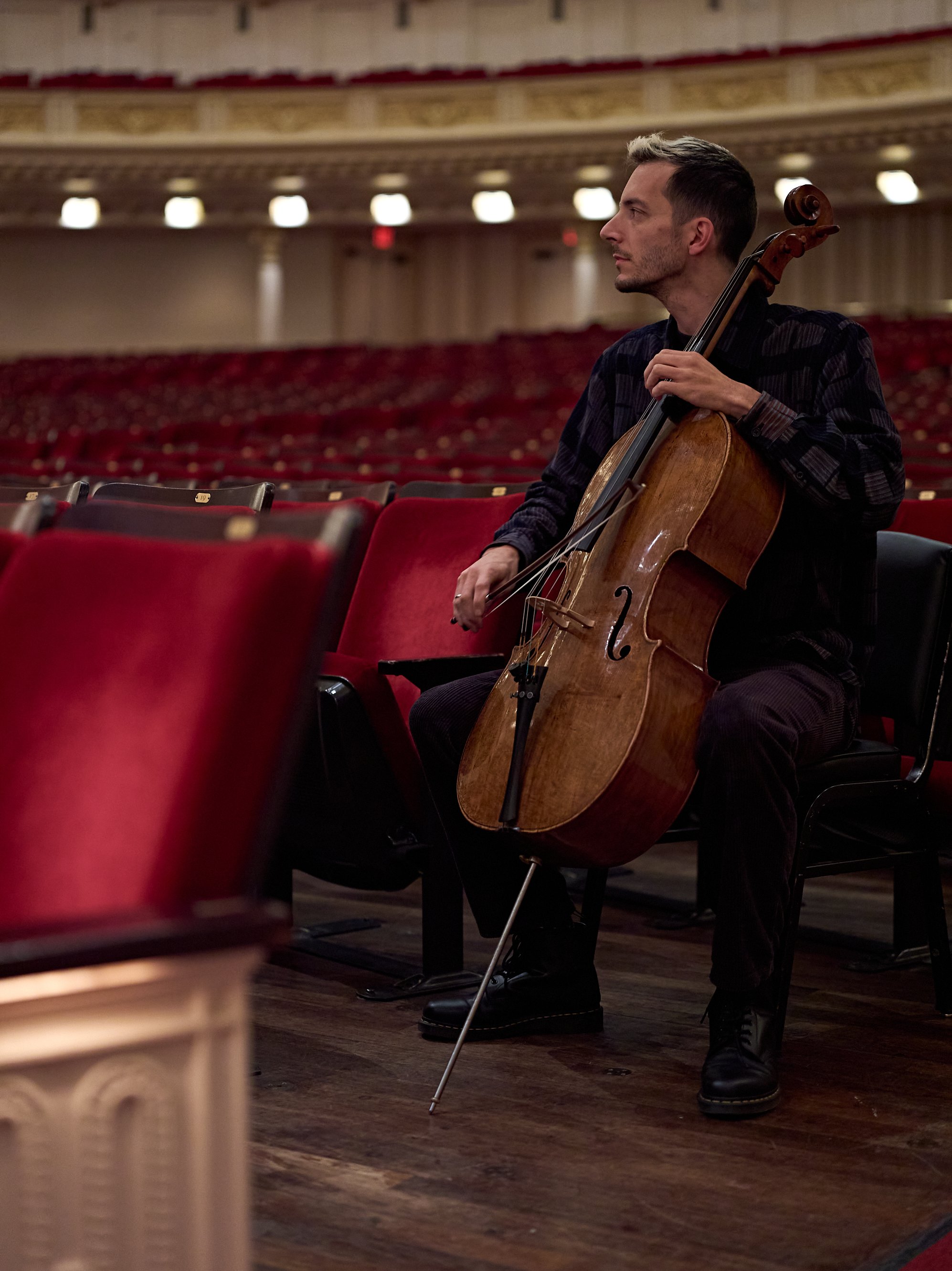 Ein Cellist sitzt auf der Bühne eines großen, leeren Konzertsaals mit roten Samtsesseln und hält sein Cello und seinen Bogen in den Händen.