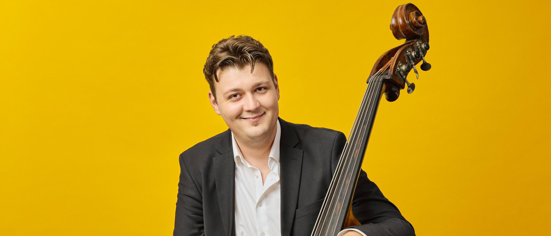 A smiling man in a suit holds a double bass and bow against a bright yellow background.