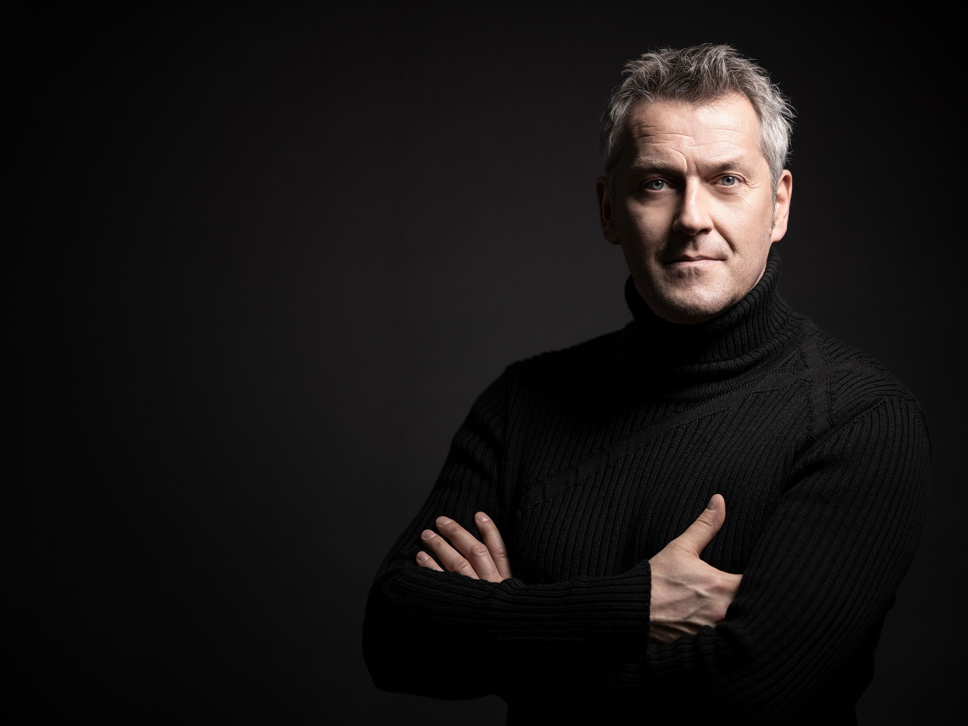 Markus Poschner mit grauem Haar und schwarzem Rollkragenpullover steht mit verschränkten Armen vor einem dunklen Hintergrund und schaut selbstbewusst in die Kamera. Die Beleuchtung hebt sein Gesicht und seinen Oberkörper hervor.