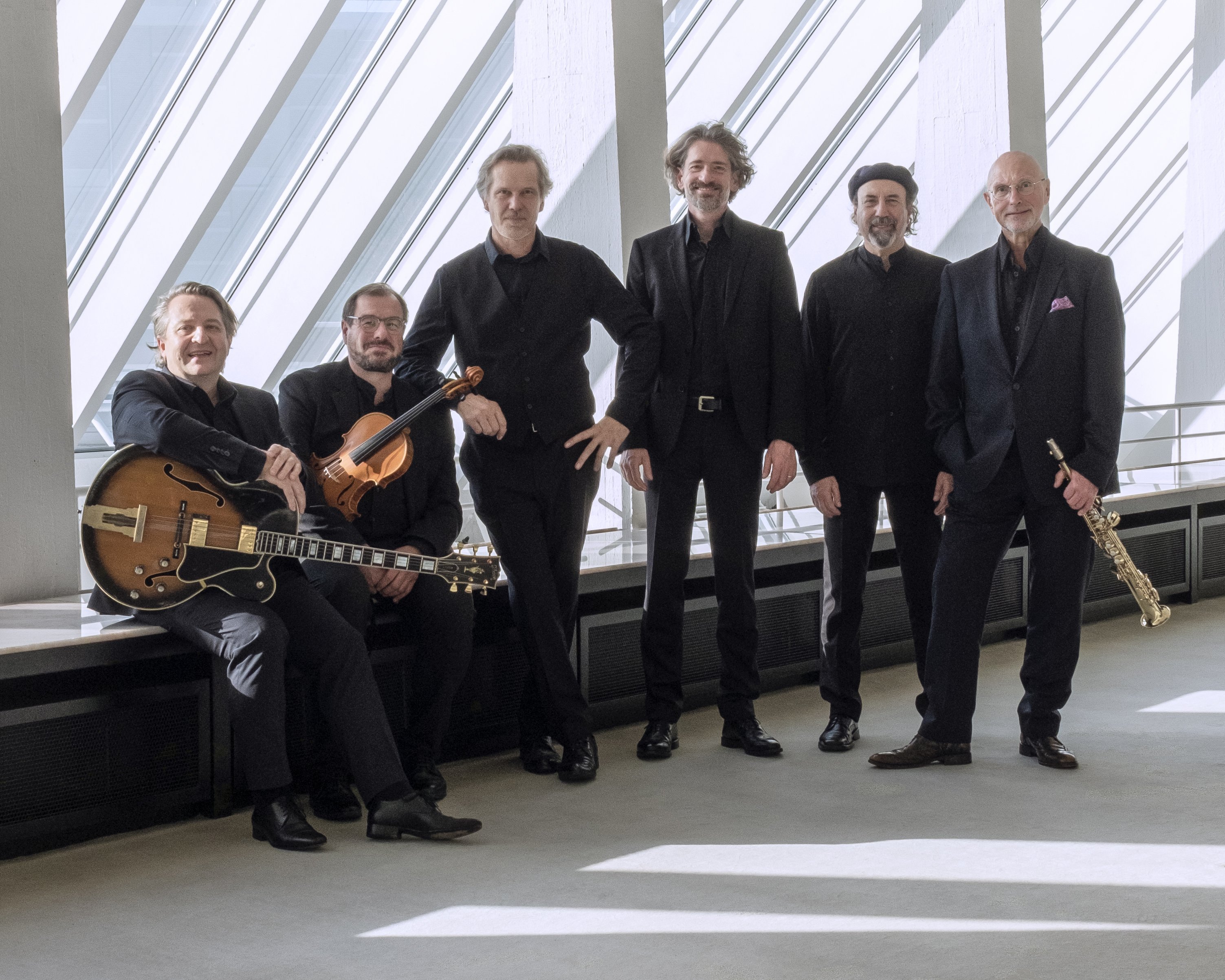 Sechs schwarz gekleidete Männer posieren in einem Innenraum vor großen Fenstern mit schrägen Balken. Einer hält eine Gitarre, ein anderer eine Geige und ein weiterer ein Saxophon; die anderen stehen oder sitzen und lächeln in die Kamera.