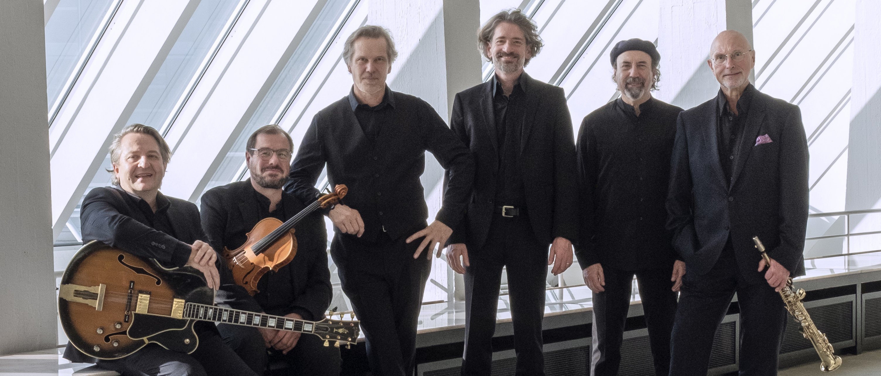 Sechs schwarz gekleidete Männer posieren in einem Innenraum vor großen Fenstern mit schrägen Balken. Einer hält eine Gitarre, ein anderer eine Geige und ein weiterer ein Saxophon; die anderen stehen oder sitzen und lächeln in die Kamera.