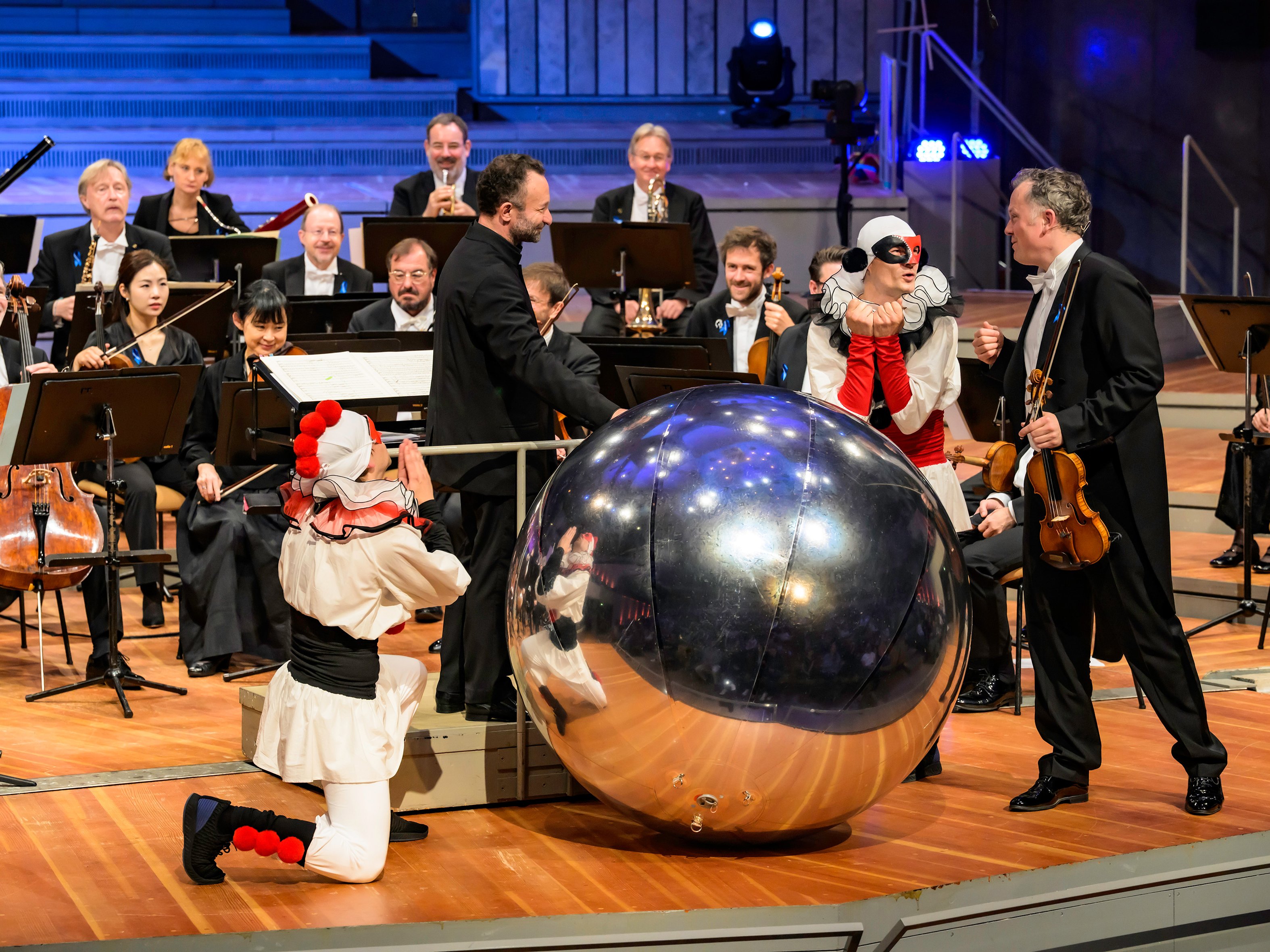 Ein Dirigent, ein Geiger und zwei Darsteller in Clownskostümen interagieren auf der Orchesterbühne mit einer großen, silbernen, spiegelnden Kugel.