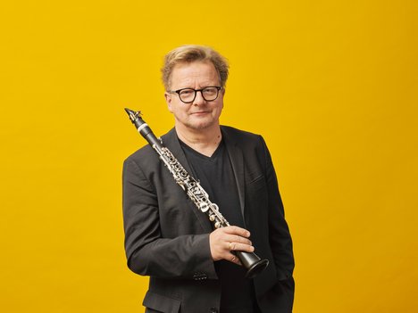 A portrait photo of Wenzel Fuchs, standind in front of a yellow background with his clarinet in his hands.