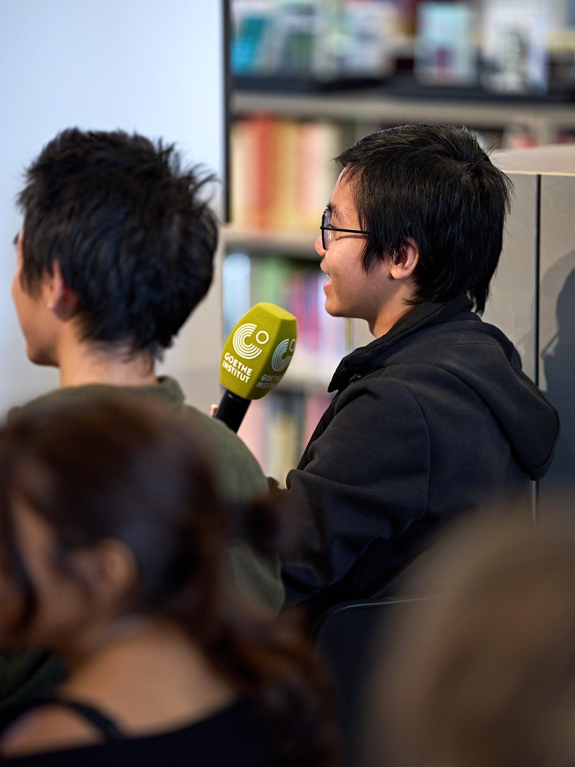 Ein junger Mann mit Brille hält ein Mikrofon des Goethe-Instituts in der Hand, während er bei einer Veranstaltung in einer Bibliothek oder einer Buchhandlung spricht.