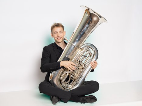 Jannik Schmidt in a black suit sits cross-legged on the floor, smiling and holding a large silver tuba against a plain white background.