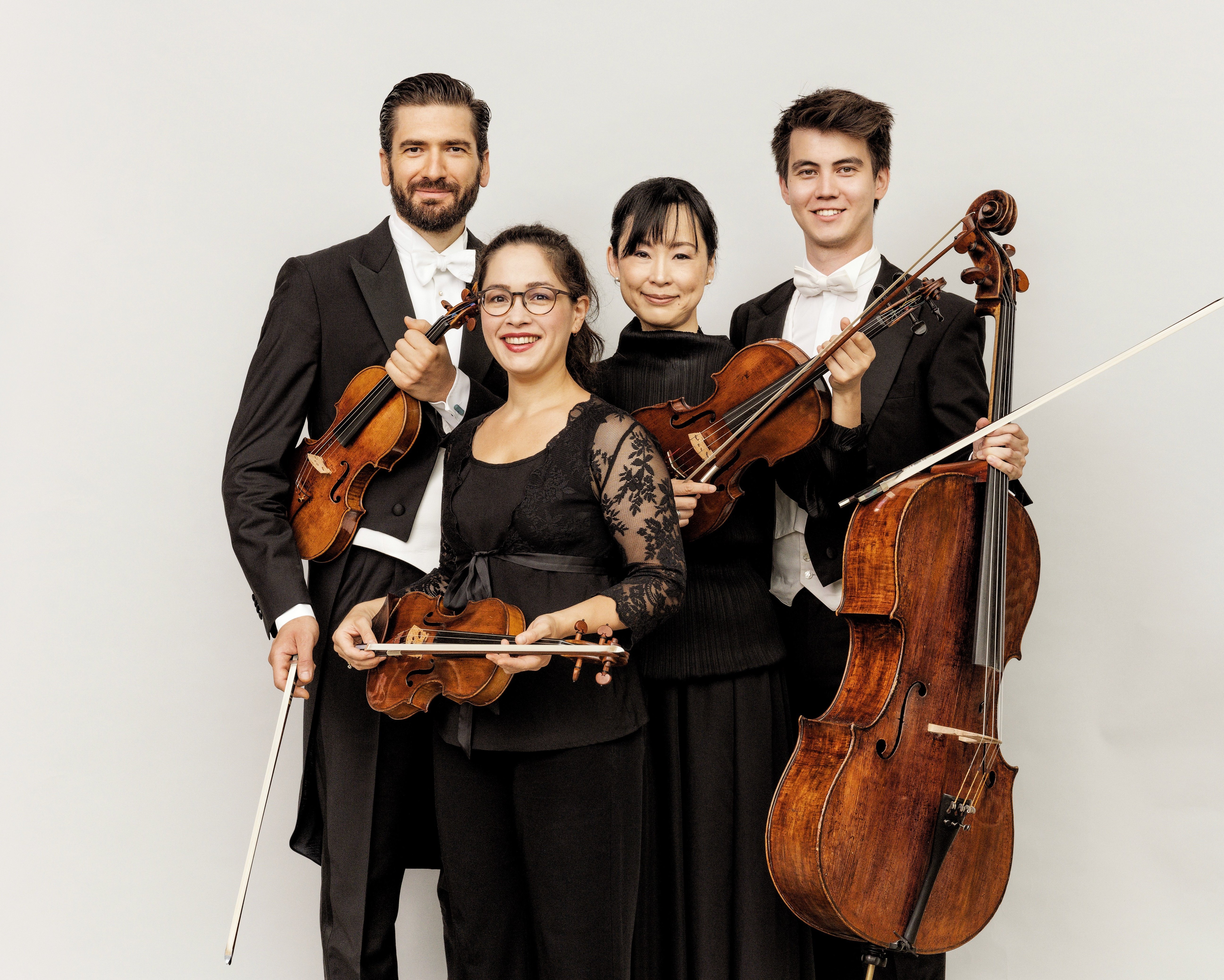 Vier klassische Musiker posieren zusammen und lächeln. Zwei halten Geigen, einer eine Bratsche und einer ein Cello. Sie tragen formelle schwarze und weiße Konzertkleidung vor einem schlichten hellen Hintergrund.