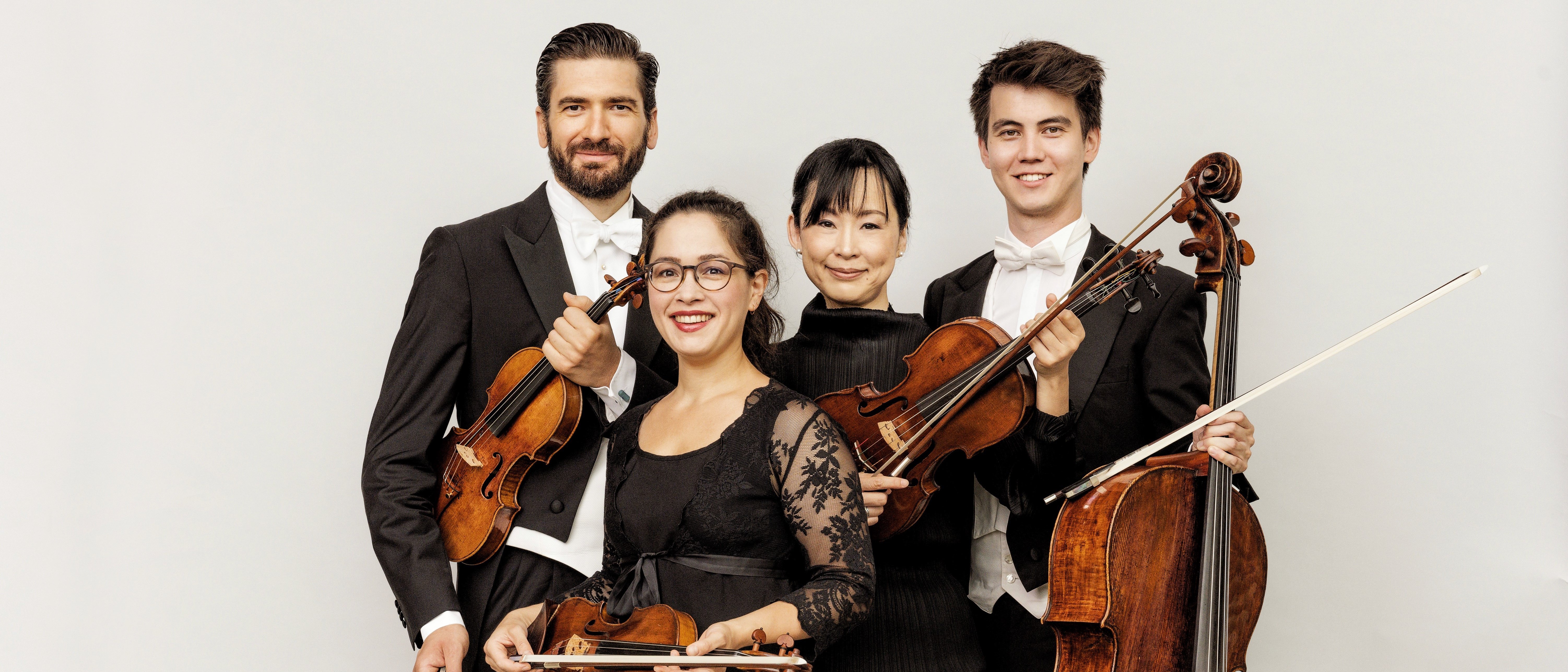 Vier klassische Musiker posieren zusammen und lächeln. Zwei halten Geigen, einer eine Bratsche und einer ein Cello. Sie tragen formelle schwarze und weiße Konzertkleidung vor einem schlichten hellen Hintergrund.