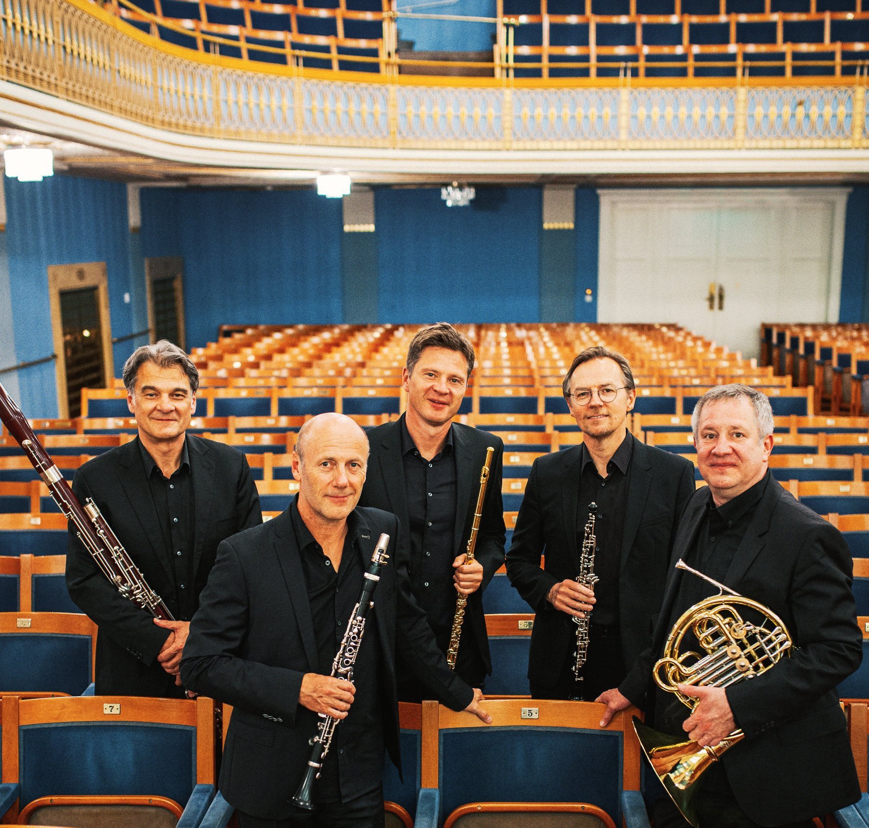 Fünf Männer in schwarzen Anzügen, die Orchesterinstrumente in der Hand halten, stehen inmitten von Reihen leerer Theatersitze und posieren für ein Gruppenfoto in einem hell erleuchteten, eleganten Konzertsaal.
