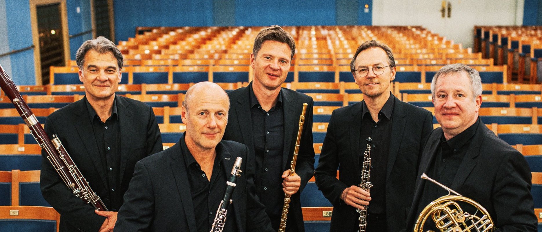 Fünf Männer in schwarzen Anzügen, die Orchesterinstrumente in der Hand halten, stehen inmitten von Reihen leerer Theatersitze und posieren für ein Gruppenfoto in einem hell erleuchteten, eleganten Konzertsaal.
