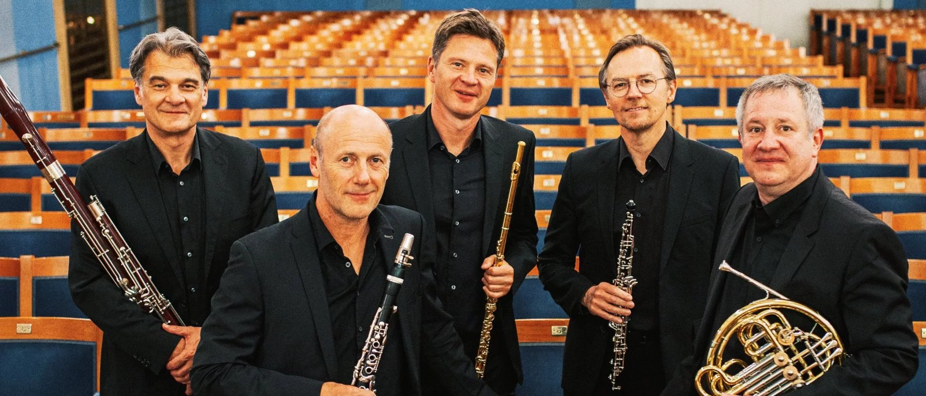 Five men in black suits holding wind instruments stand among empty auditorium seats, posing for a group photo in a bright, elegant concert hall with blue and gold accents.