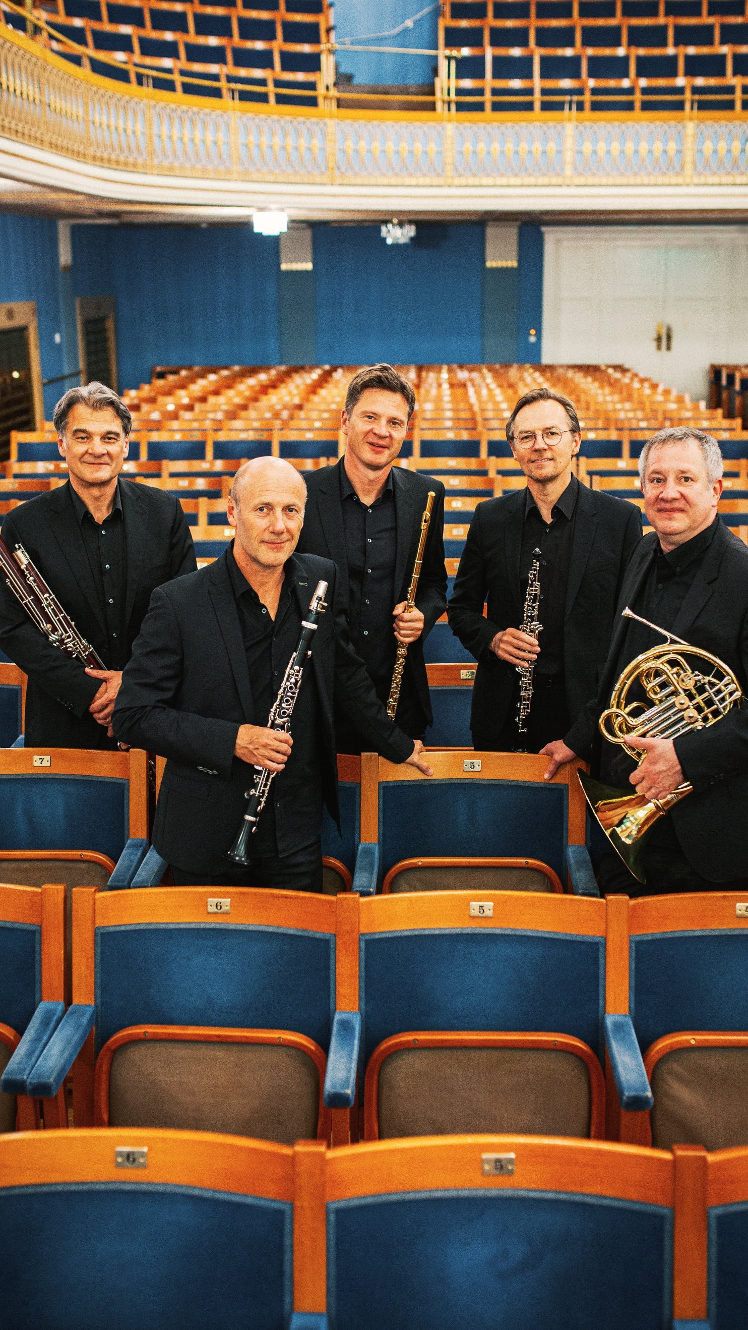 Fünf Männer in schwarzen Anzügen, die Orchesterinstrumente in der Hand halten, stehen inmitten von Reihen leerer Theatersitze und posieren für ein Gruppenfoto in einem hell erleuchteten, eleganten Konzertsaal.