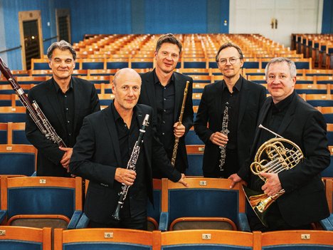 Fünf Männer in schwarzen Anzügen, die Orchesterinstrumente in der Hand halten, stehen inmitten von Reihen leerer Theatersitze und posieren für ein Gruppenfoto in einem hell erleuchteten, eleganten Konzertsaal.