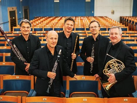 Five men in black suits holding wind instruments stand among empty auditorium seats, posing for a group photo in a bright, elegant concert hall with blue and gold accents.