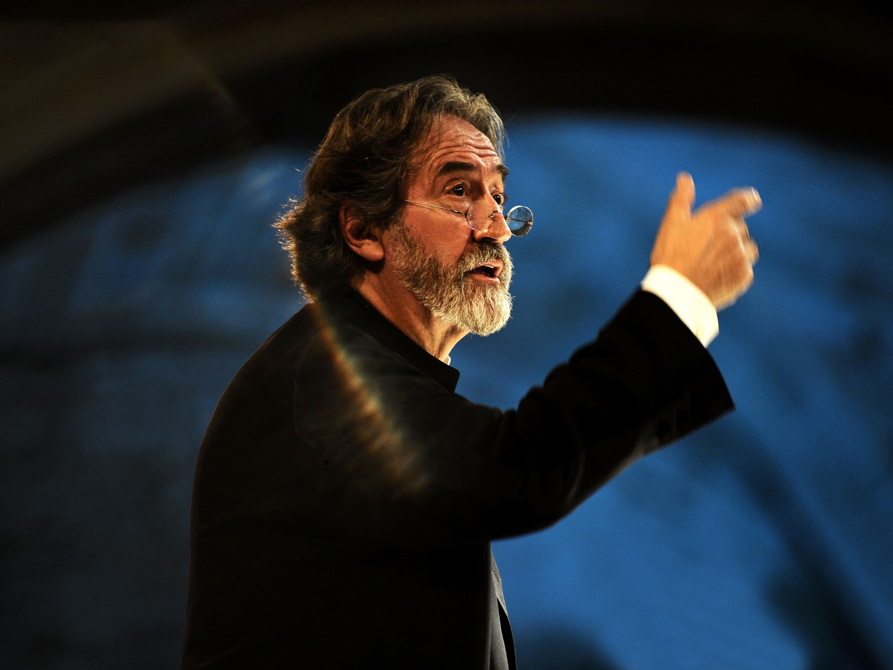 Jordi Savall mit grauem Haar und Bart gestikuliert leidenschaftlich. Er trägt eine Brille und einen schwarzen Anzug. Er tritt vor einem unscharfen Hintergrund auf, der von dramatischer Beleuchtung angestrahlt wird, die einen Regenbogeneffekt erzeugt.