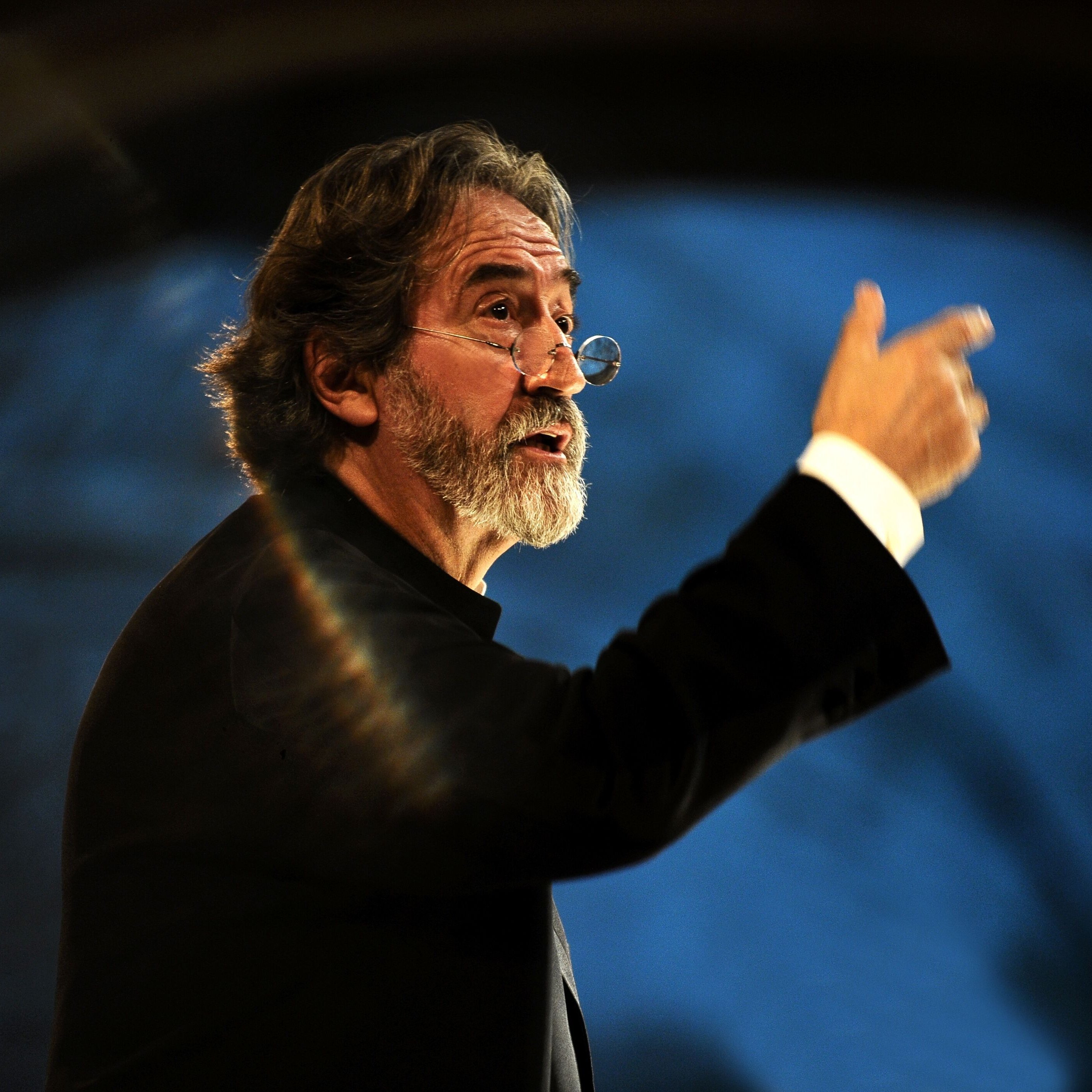 Jordi Savall, with grey hair and beard, gesticulates passionately. He wears glasses and a black suit. He appears in front of a blurred background illuminated by dramatic lighting that creates a rainbow effect.