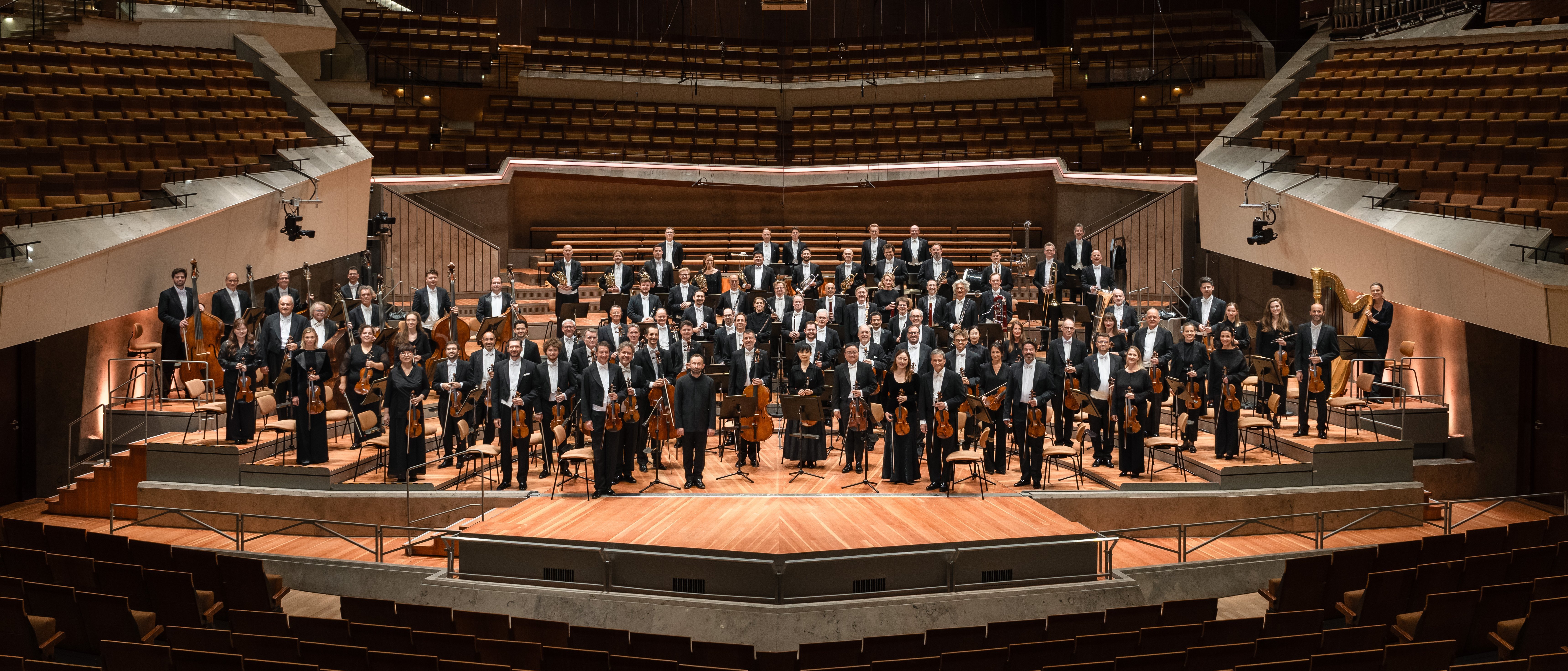 Ein formell gekleidetes Symphonieorchester steht auf der Bühne eines großen, modernen Konzertsaals mit gestufter Bestuhlung und hölzernem Interieur.