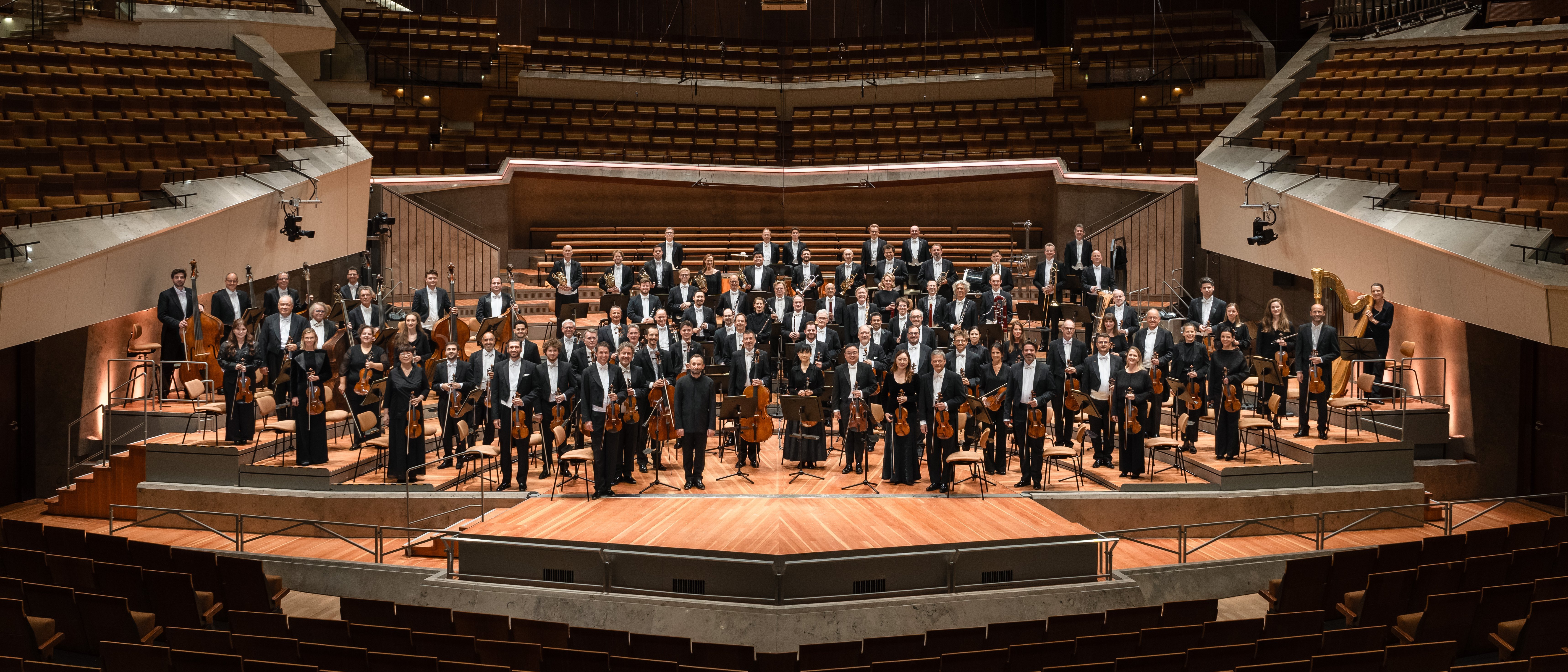Ein formell gekleidetes Symphonieorchester steht auf der Bühne eines großen, modernen Konzertsaals mit gestufter Bestuhlung und hölzernem Interieur.