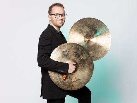 A smiling man in a black suit holds two large brass cymbals, standing against a plain white background.