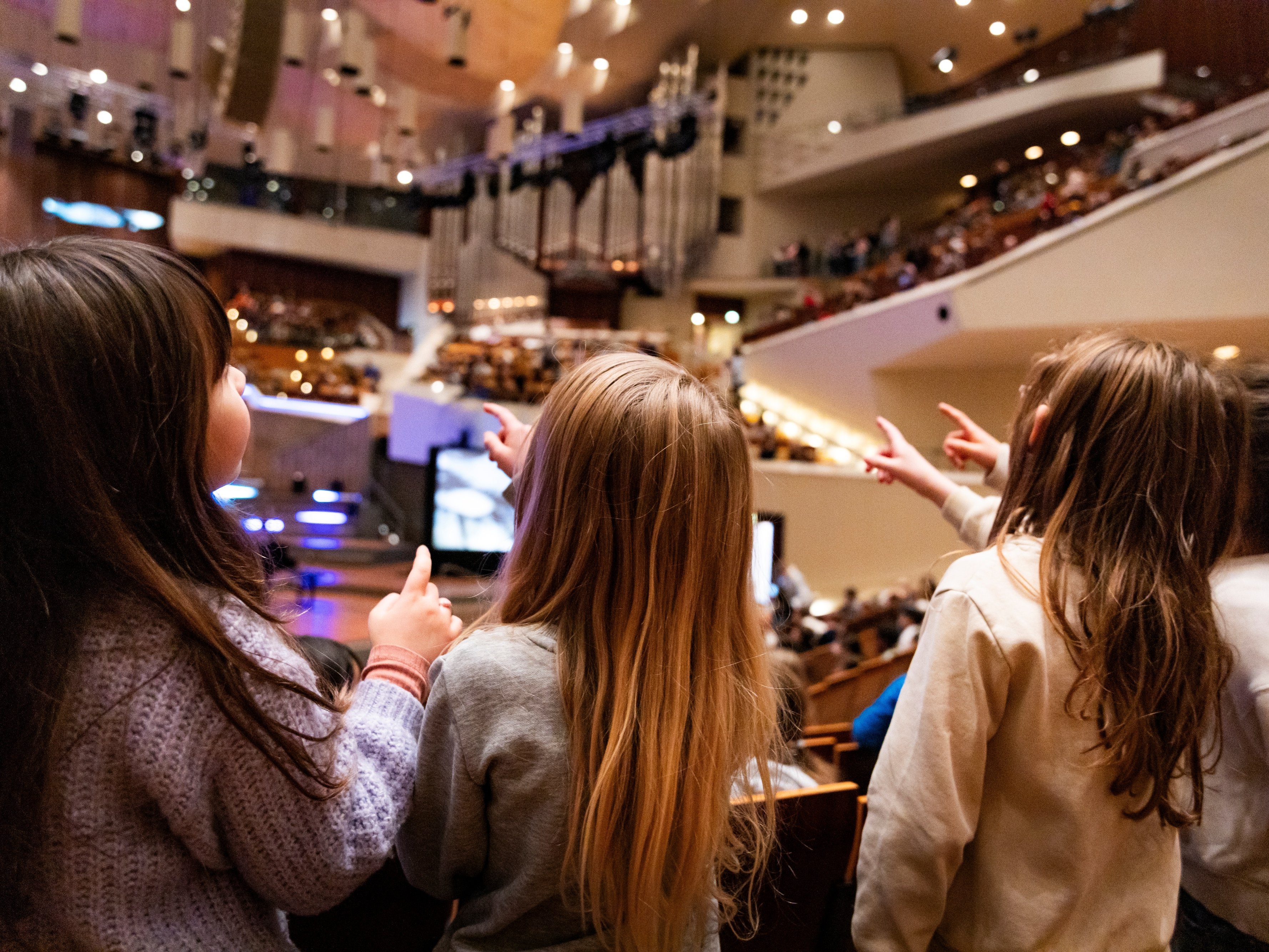 Vier Kinder mit langen Haaren, von hinten gesehen, sitzen in einem Zuschauerraum und zeigen aufgeregt auf die hell erleuchtete Bühne, auf der eine Aufführung stattzufinden scheint. Das Publikum füllt den Hintergrund.