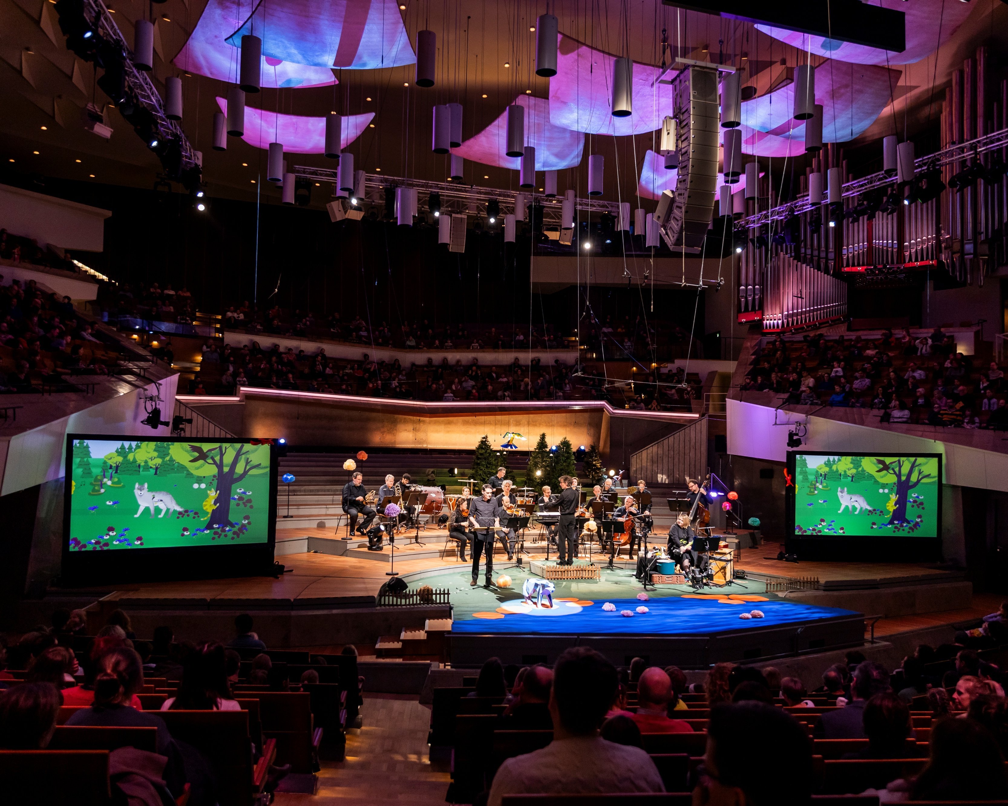 A symphony orchestra performs on stage in a concert hall, with colorful forest-themed animations displayed on two large screens and an audience seated throughout the venue.