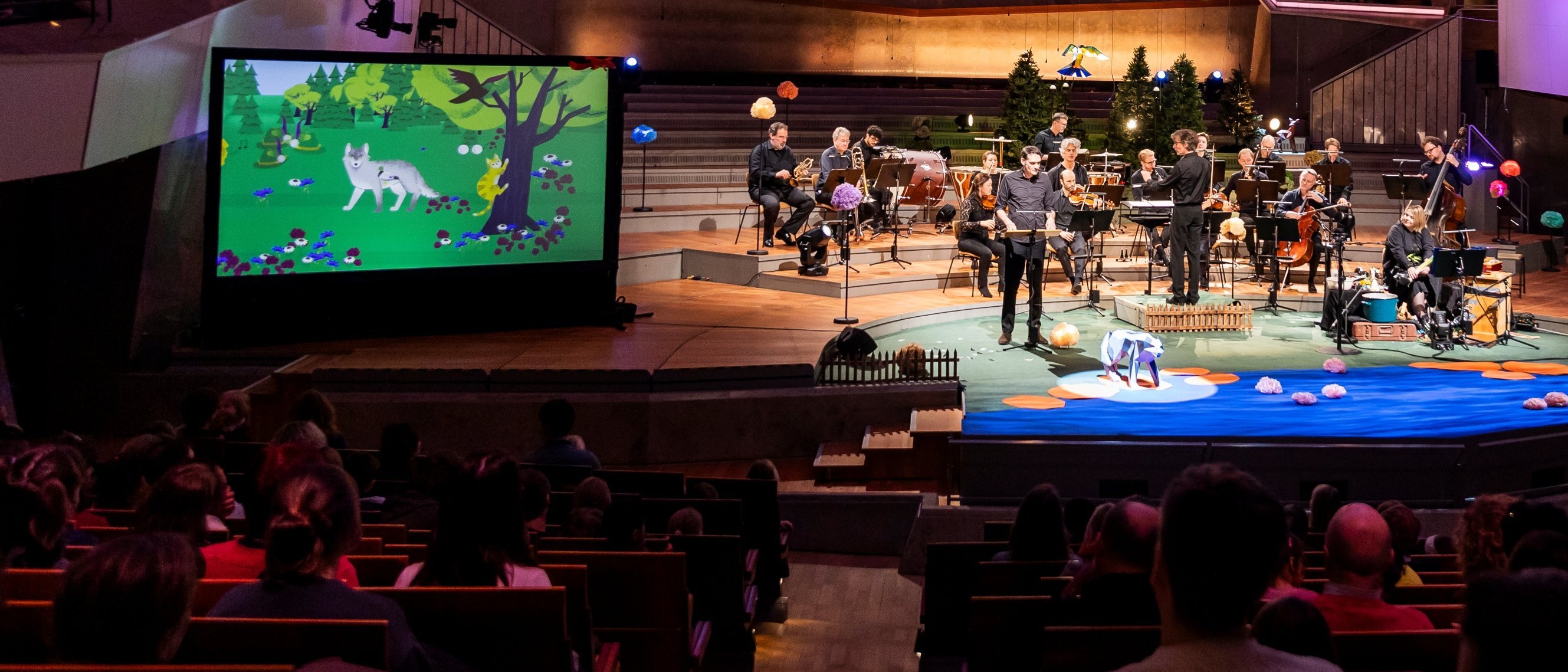 A symphony orchestra performs on stage in a concert hall, with colorful forest-themed animations displayed on two large screens and an audience seated throughout the venue.