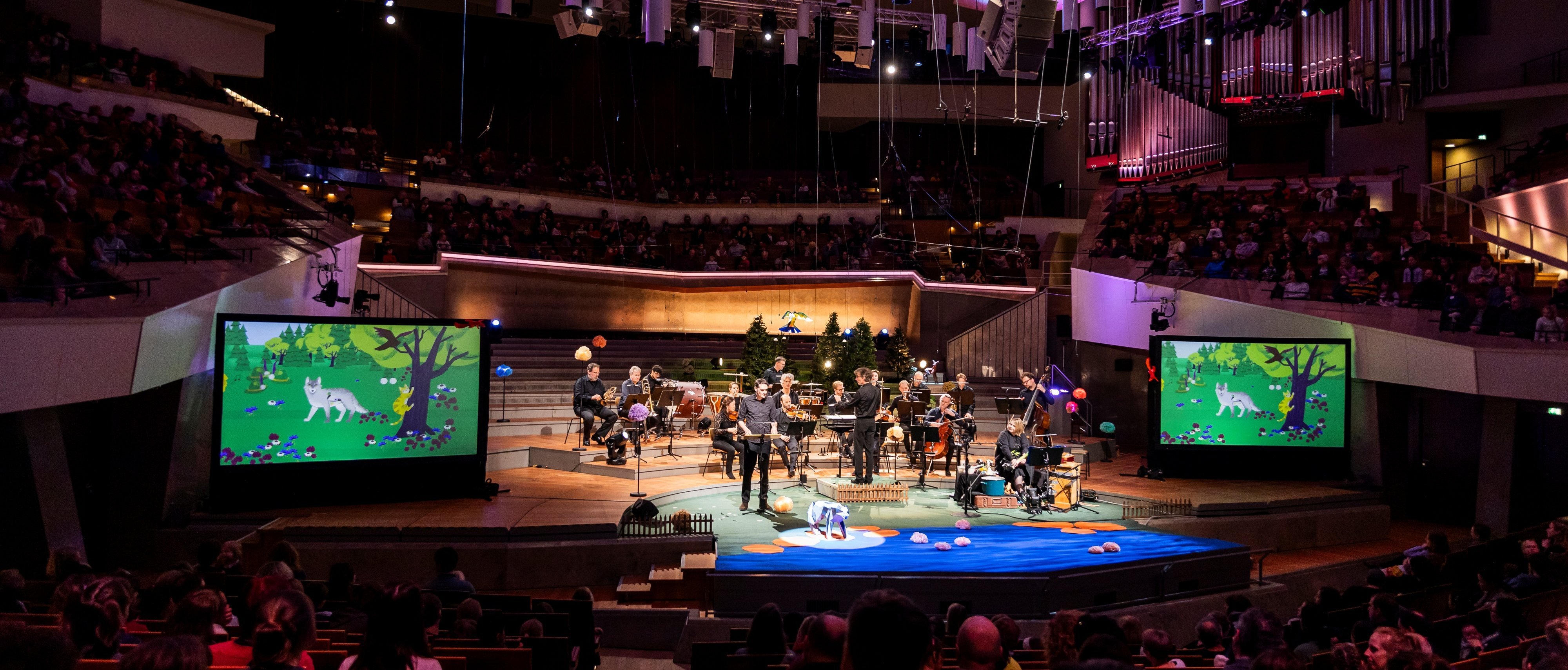 Ein Orchester spielt auf einer hell erleuchteten Bühne in einem großen Konzertsaal, in dem das Publikum sitzt. Zwei große Bildschirme zeigen eine bunte, animierte Waldszene mit Tieren auf beiden Seiten der Bühne.