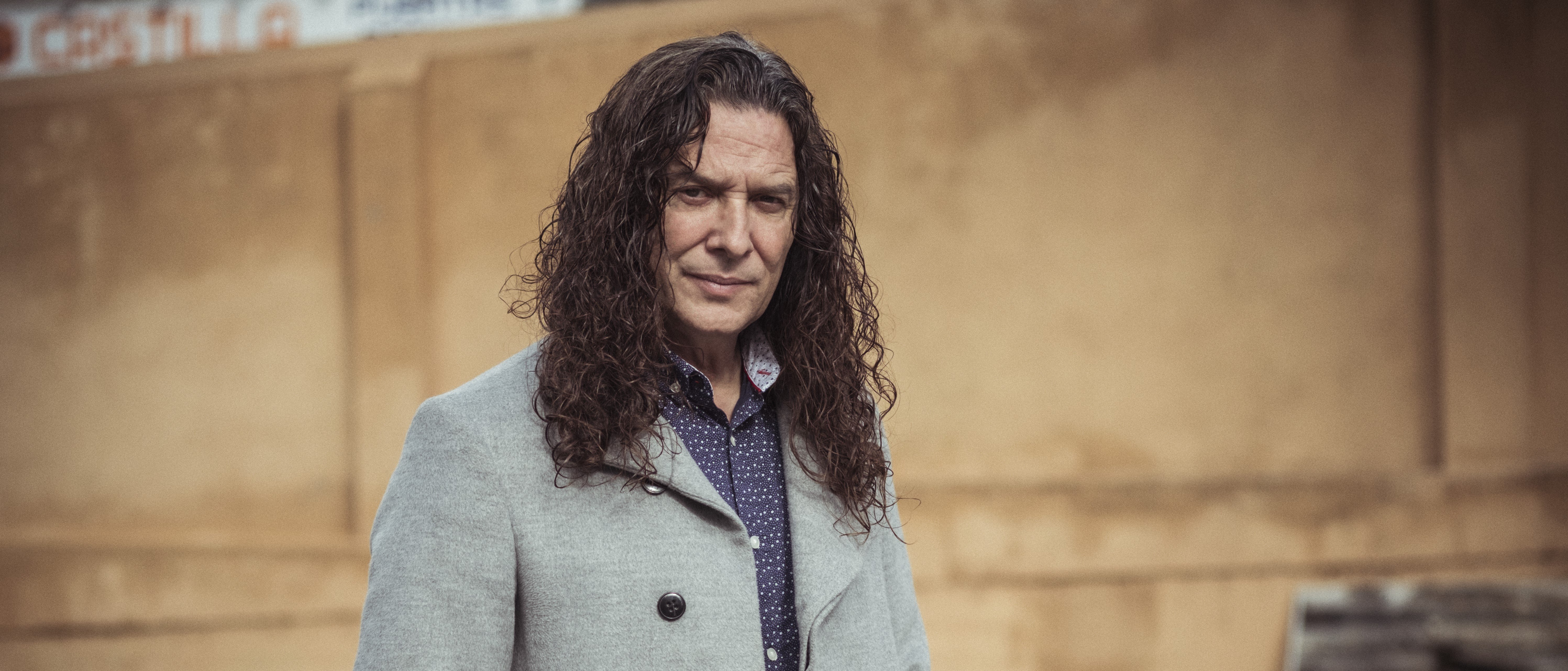 Tomatito with long curly hair, a grey coat and a polka-dot shirt is standing outside. The background is blurred and shows a beige wall and a gravel path.