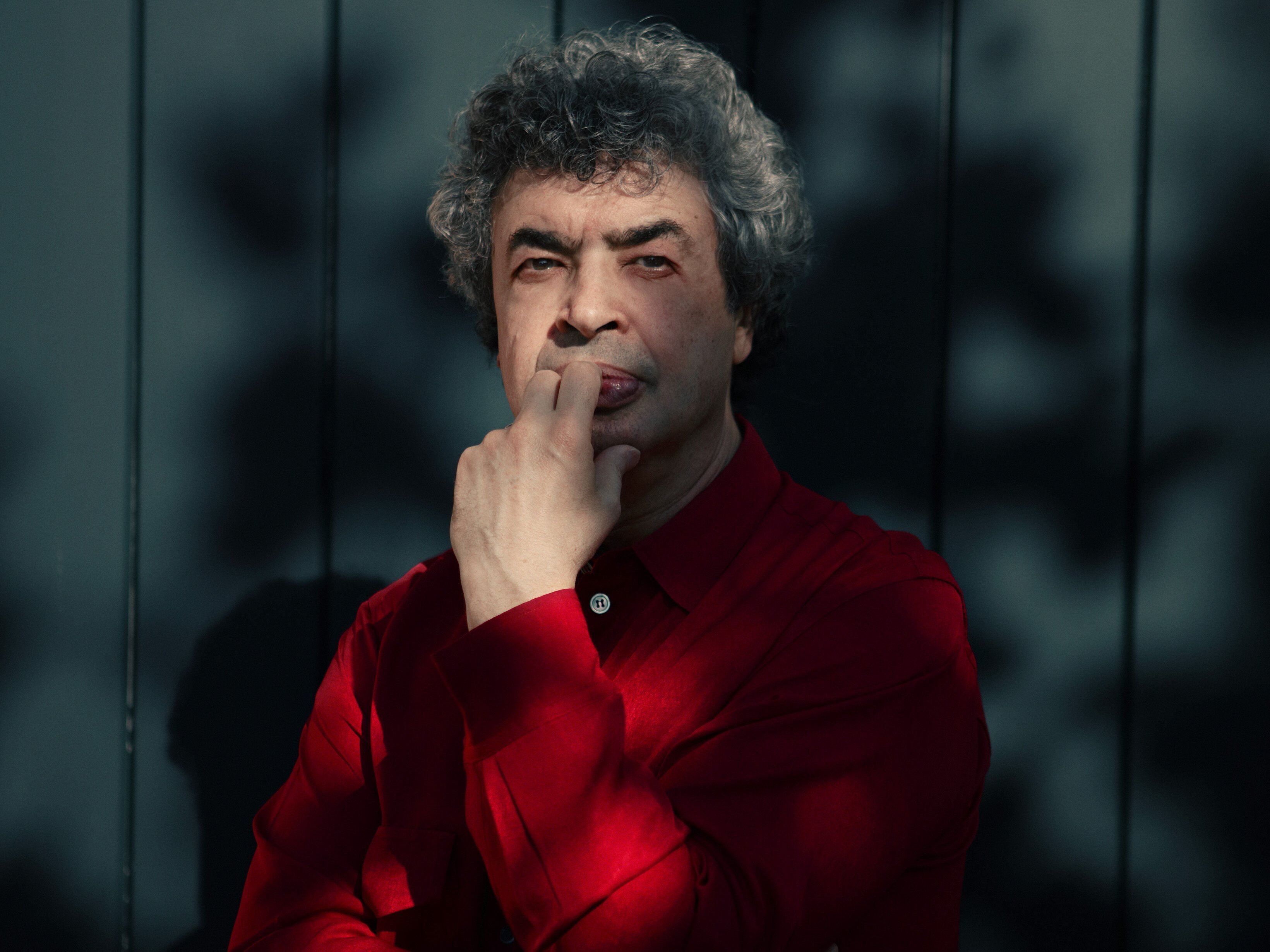 Semyon Bychkov mit lockigem Haar und einem roten Hemd steht nachdenklich vor einem dunklen Hintergrund mit Baumschatten. Sie hat eine Hand am Kinn und die andere vor der Brust verschränkt.