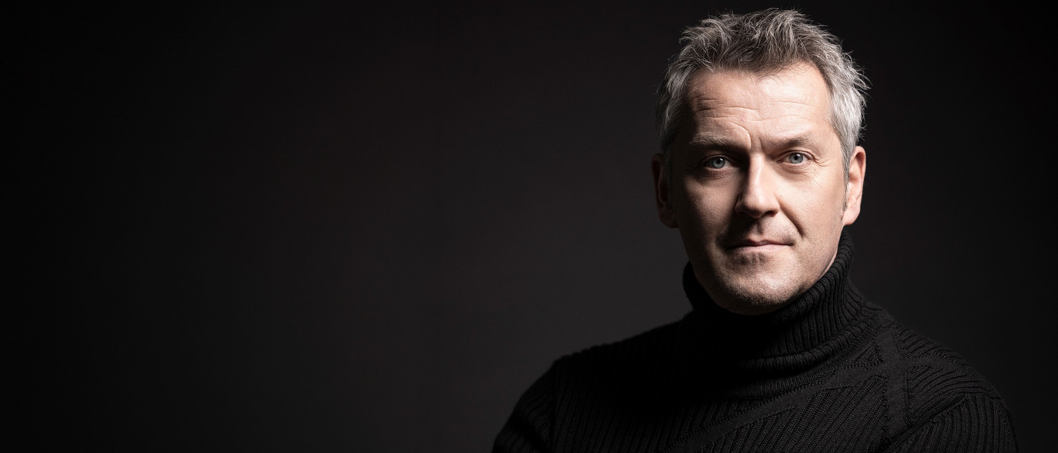 Markus Poschner mit grauem Haar und schwarzem Rollkragenpullover steht mit verschränkten Armen vor einem dunklen Hintergrund und schaut selbstbewusst in die Kamera. Die Beleuchtung hebt sein Gesicht und seinen Oberkörper hervor.