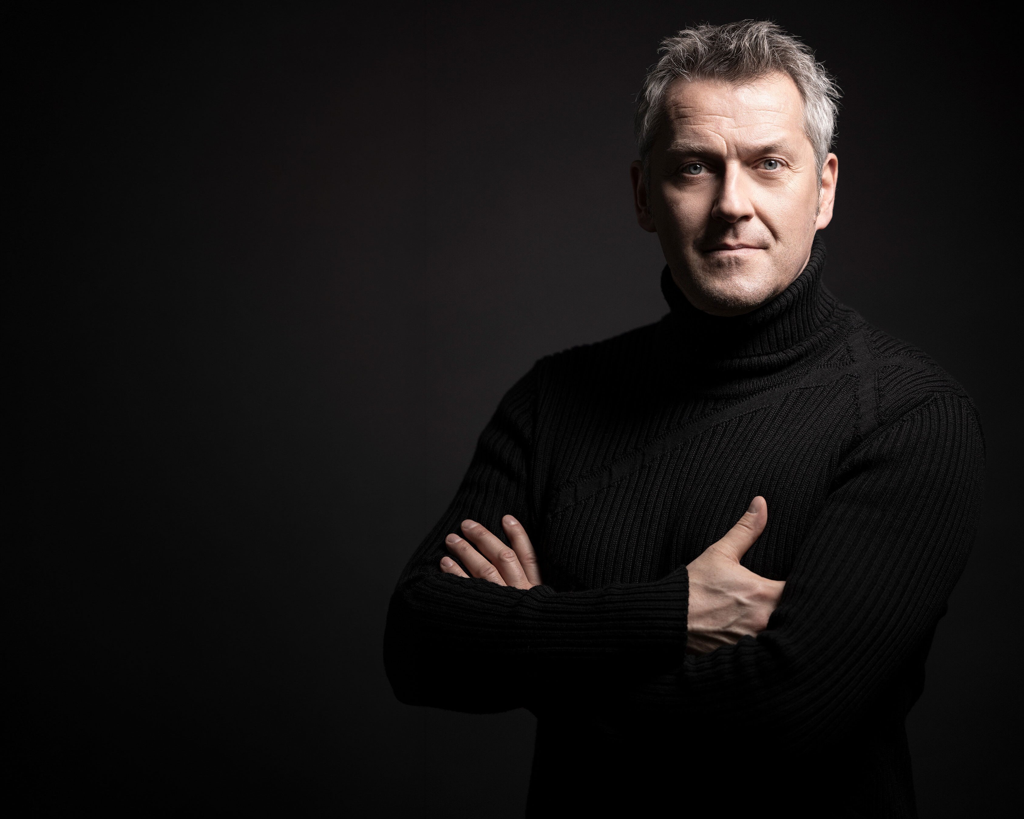Markus Poschner mit grauem Haar und schwarzem Rollkragenpullover steht mit verschränkten Armen vor einem dunklen Hintergrund und schaut selbstbewusst in die Kamera. Die Beleuchtung hebt sein Gesicht und seinen Oberkörper hervor.