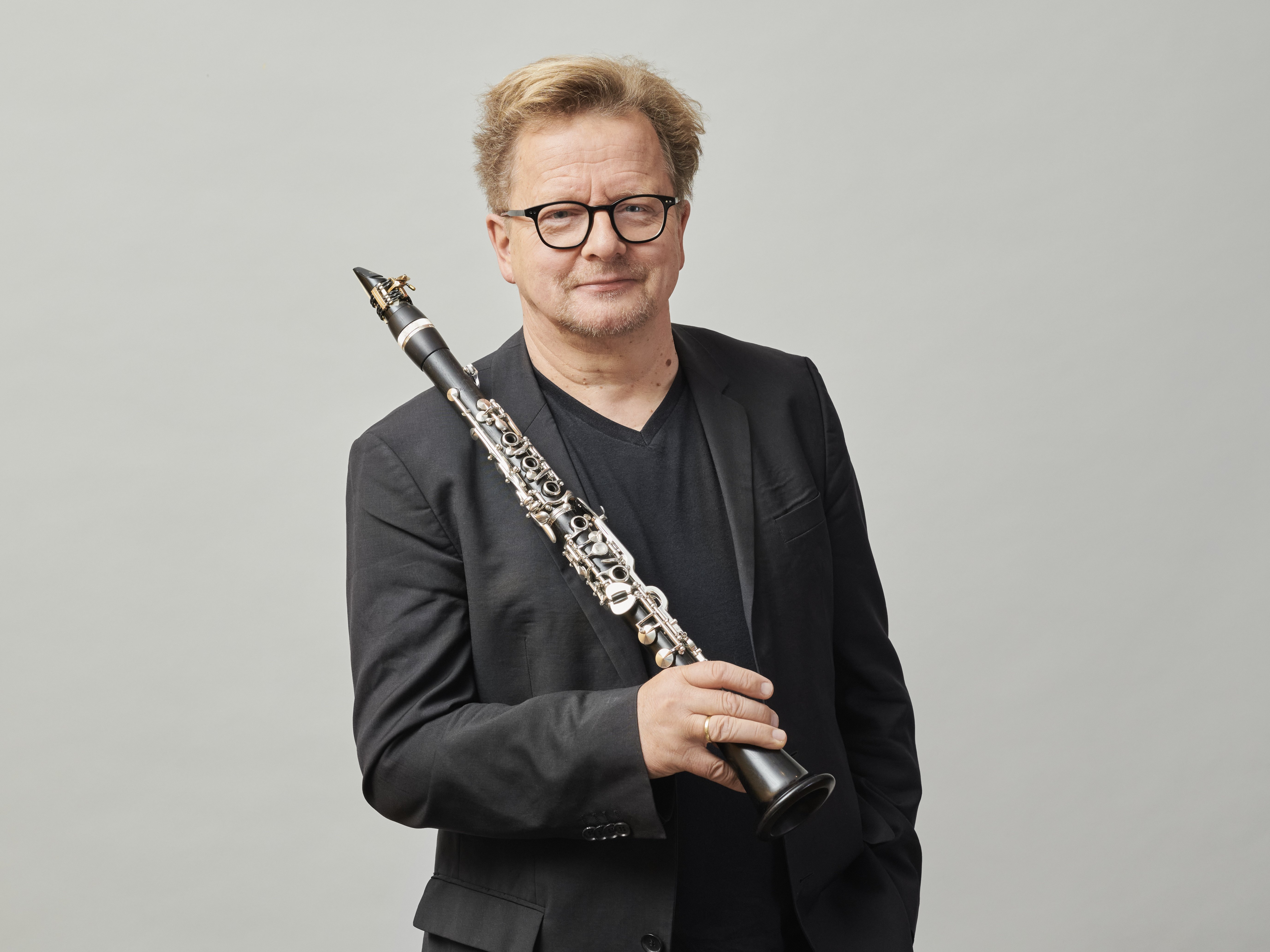 Wenzel Fuchs mit kurzen, hellen Haaren und Brille, der einen schwarzen Blazer und ein schwarzes Hemd trägt, steht vor einem schlichten Hintergrund und hält eine Klarinette aufrecht in seiner rechten Hand.