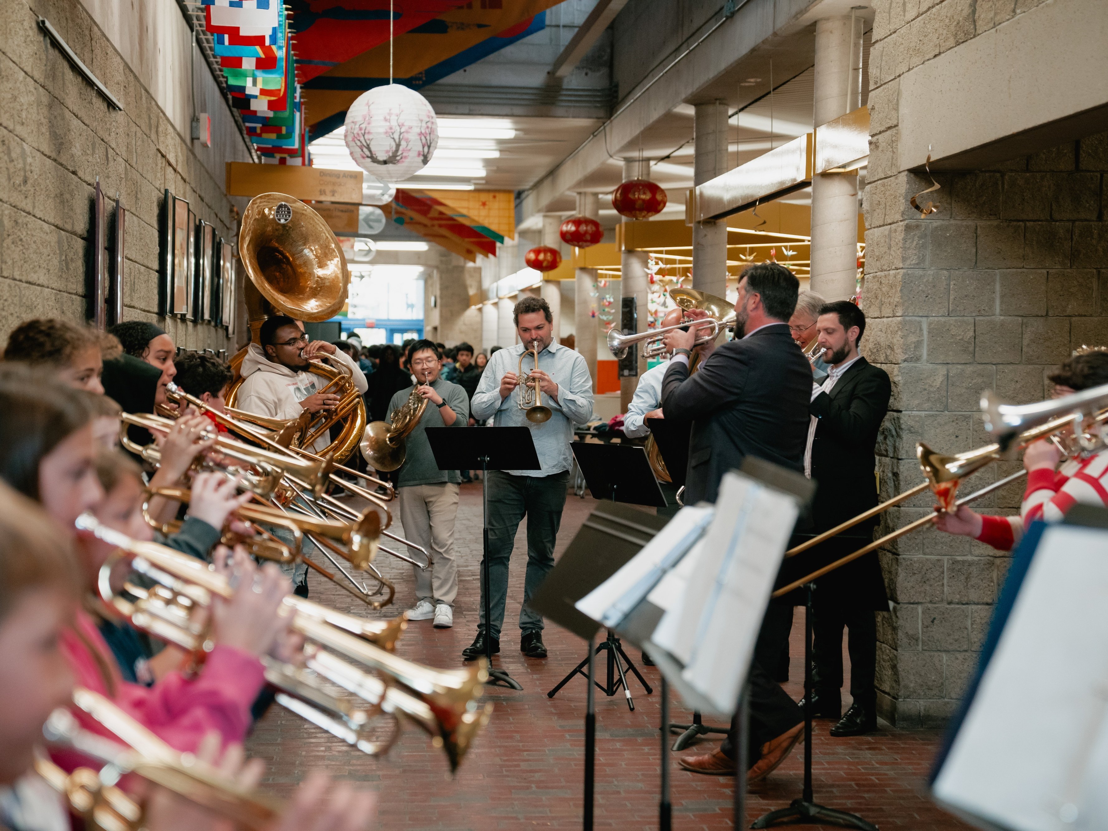 Eine große Gruppe von Musikern spielt auf Blechblasinstrumenten in einem mit Laternen und bunten Bannern geschmückten Flur, während ein Dirigent sie leitet.