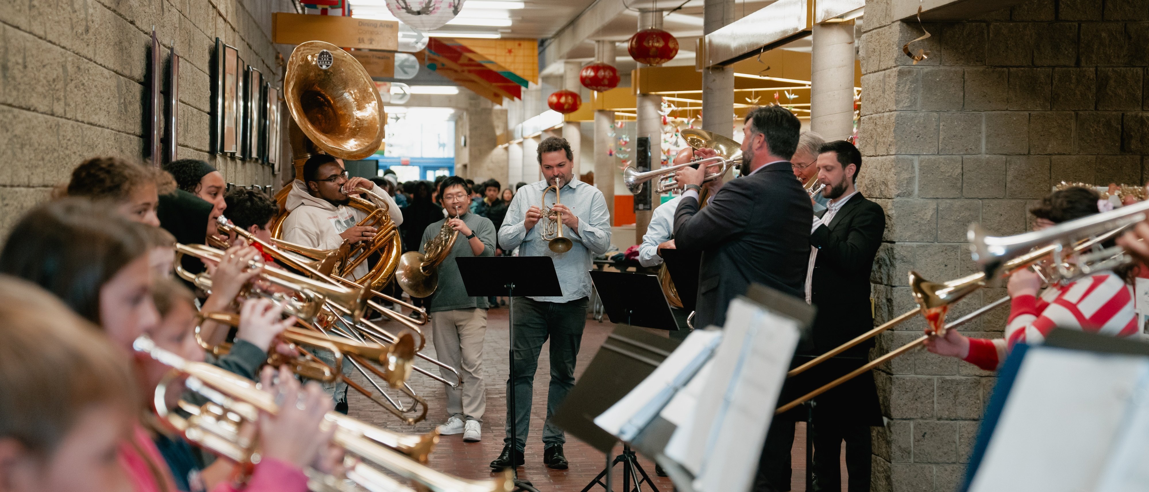 Eine große Gruppe von Musikern spielt auf Blechblasinstrumenten in einem mit Laternen und bunten Bannern geschmückten Flur, während ein Dirigent sie leitet.