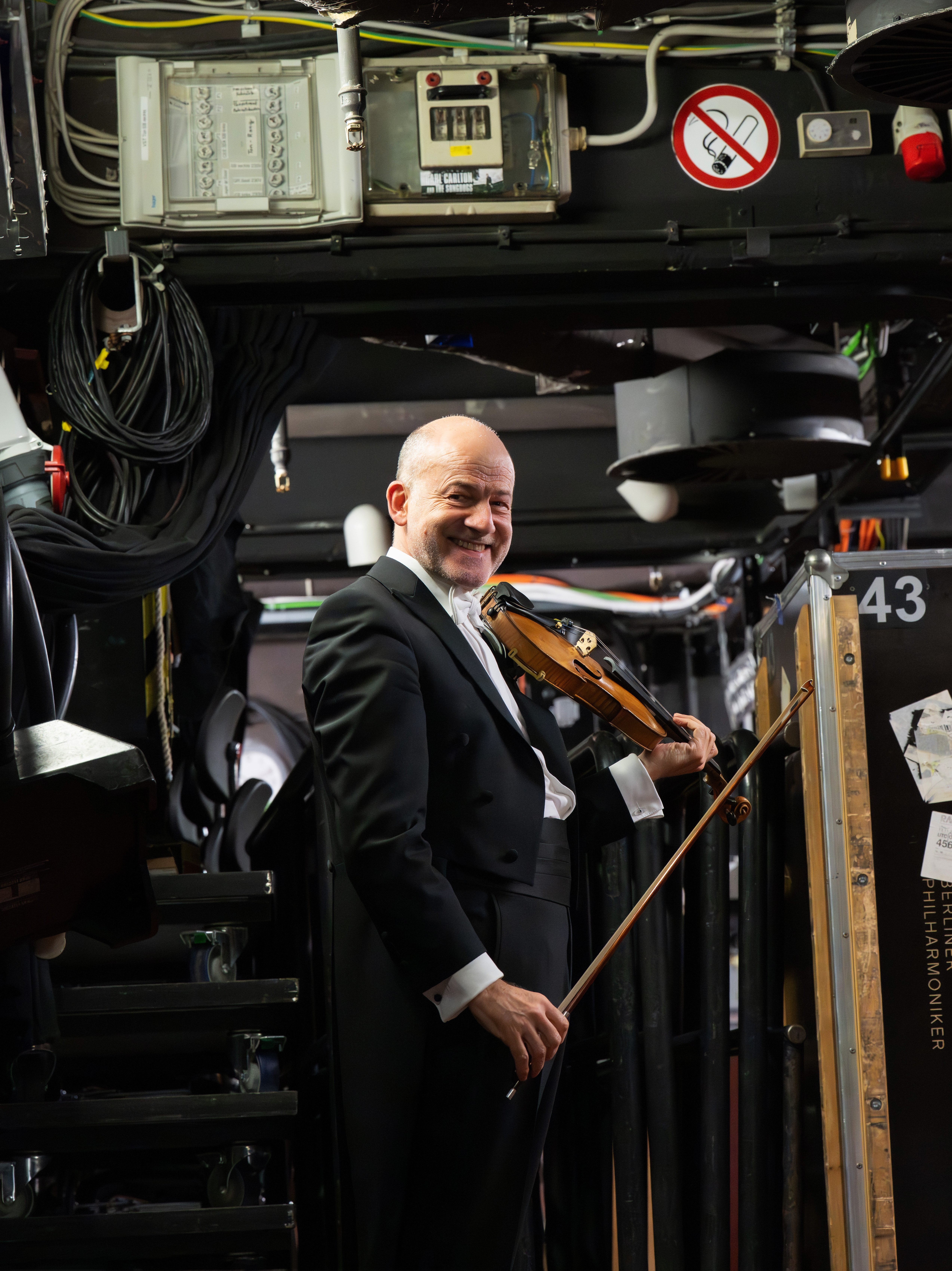 Christoph Streuli in the backstage area, surrounded by pipes, cables, and equipment. No smoking signs and industrial fixtures are visible above him.