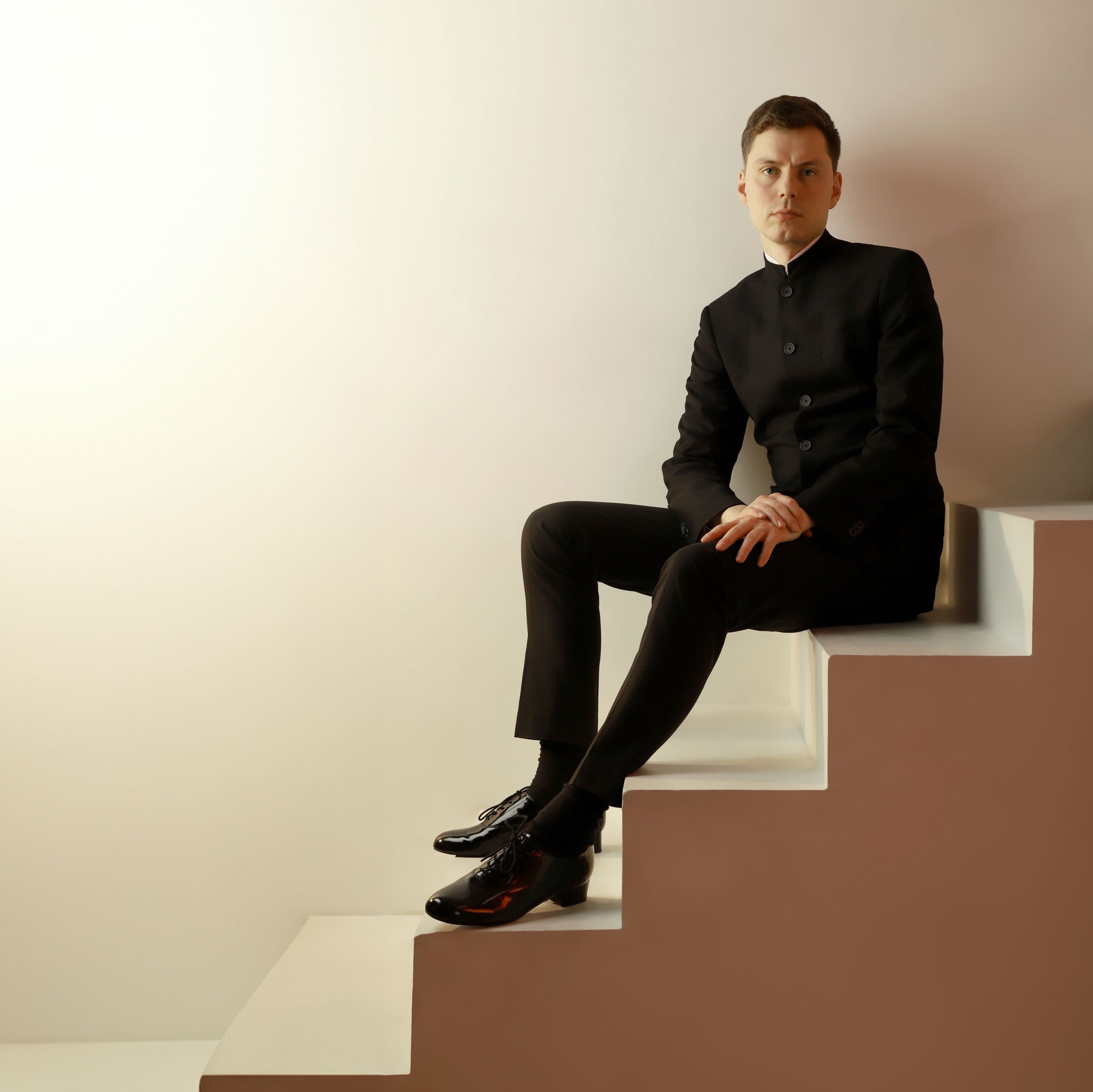 Richard Gowers im schwarzen Anzug sitzt auf einer minimalistischen, blockartigen Treppe vor einem schlichten Hintergrund mit sanftem Hell-Dunkel-Farbverlauf. Die Hände ruhen im Schoß, der Blick ist direkt in die Kamera gerichtet.