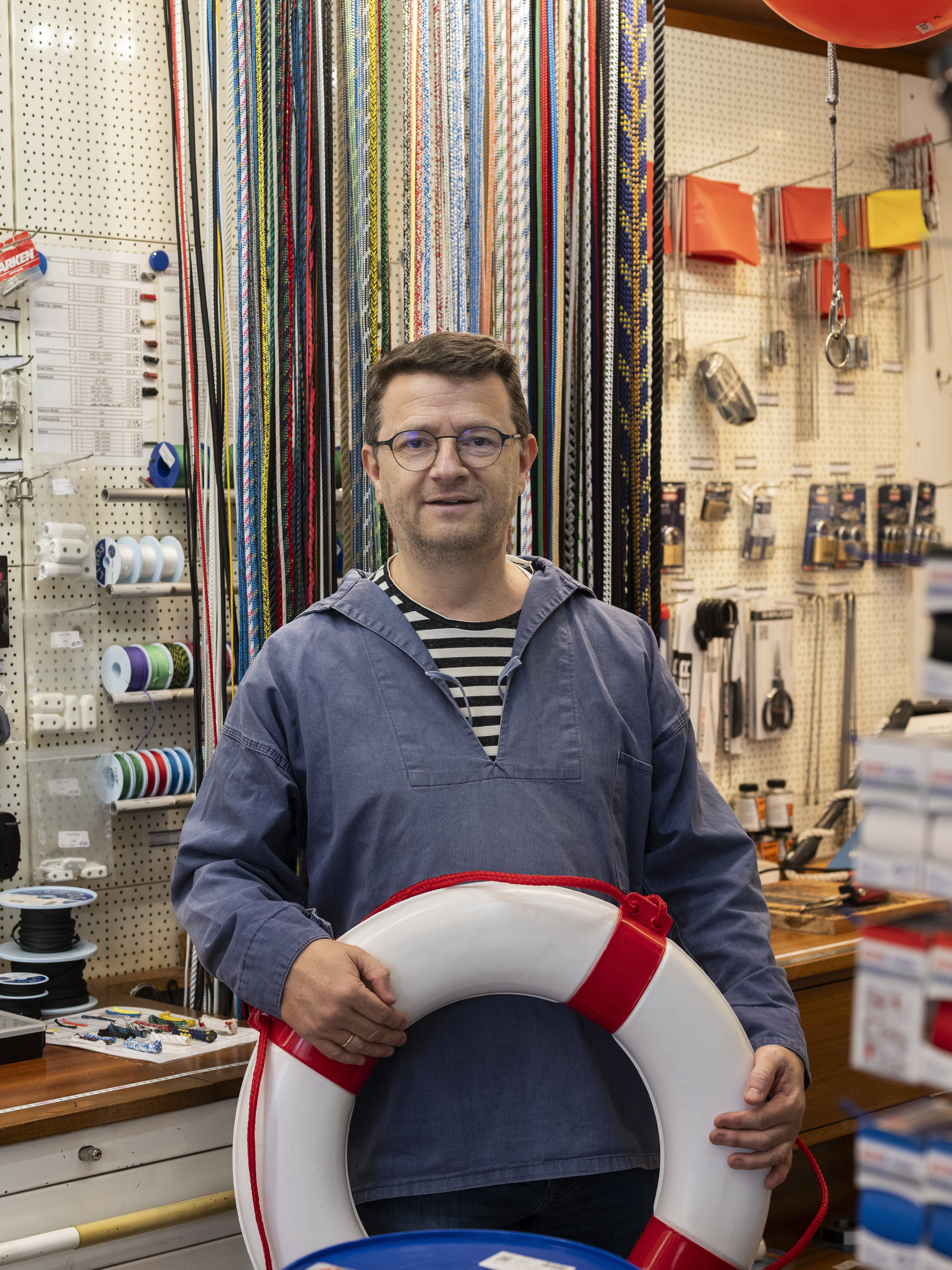 A man wearing glasses and a blue shirt stands in a hardware or marine supply store, holding a life buoy. Behind him are colorful ropes, tools, accessories, and some red balloons.