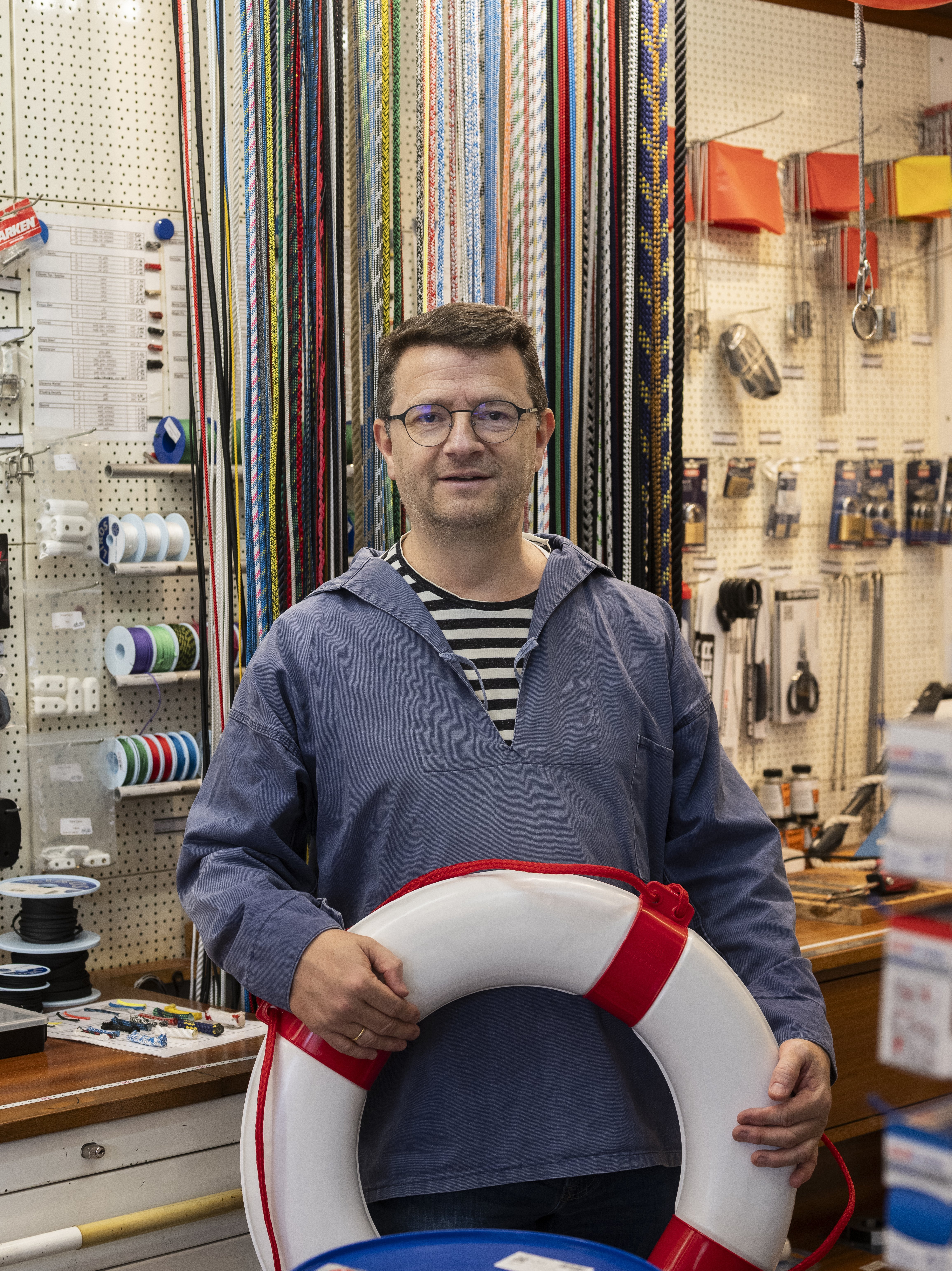 Ein Mann mit Brille und blauem Hemd hält eine Rettungsweste in der Hand. Hinter ihm sind bunte Seile, Werkzeuge und Baumaterialien zu sehen, und über ihm hängen rote Luftballons.